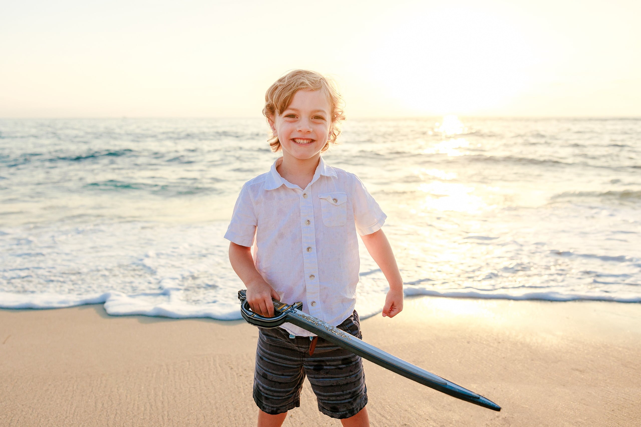 La Jolla Family Photography — 06 Child in white shirt smiling while holding pirate sword on beach at golden hour