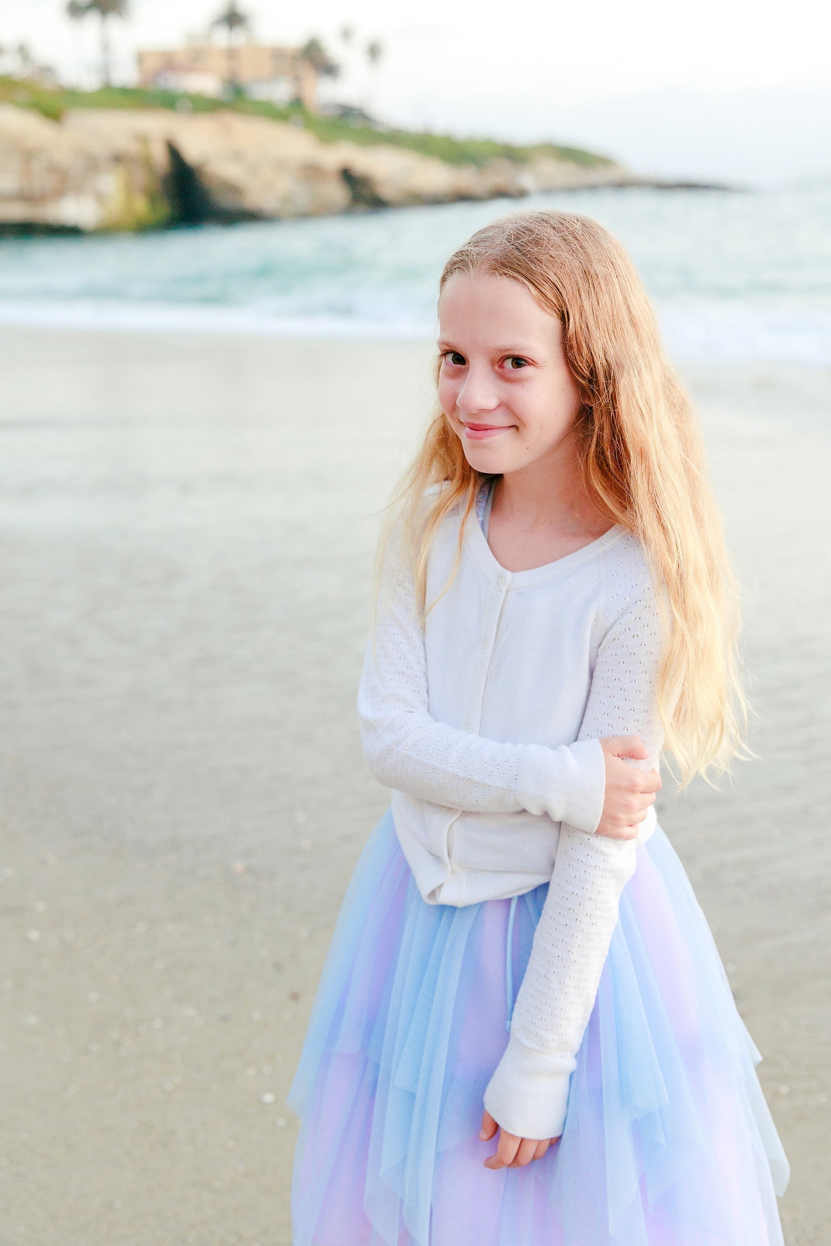 La Jolla Family Photography — 52 Girl in white cardigan and light purple tulle on beach with rocky cliff