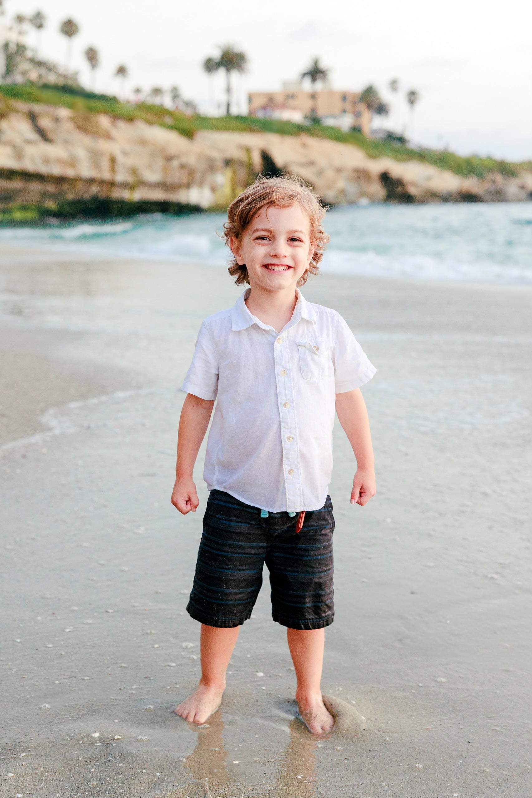 La Jolla Family Photography — 58 Toddler in white shirt and black shorts on wet sand with rocky cliff