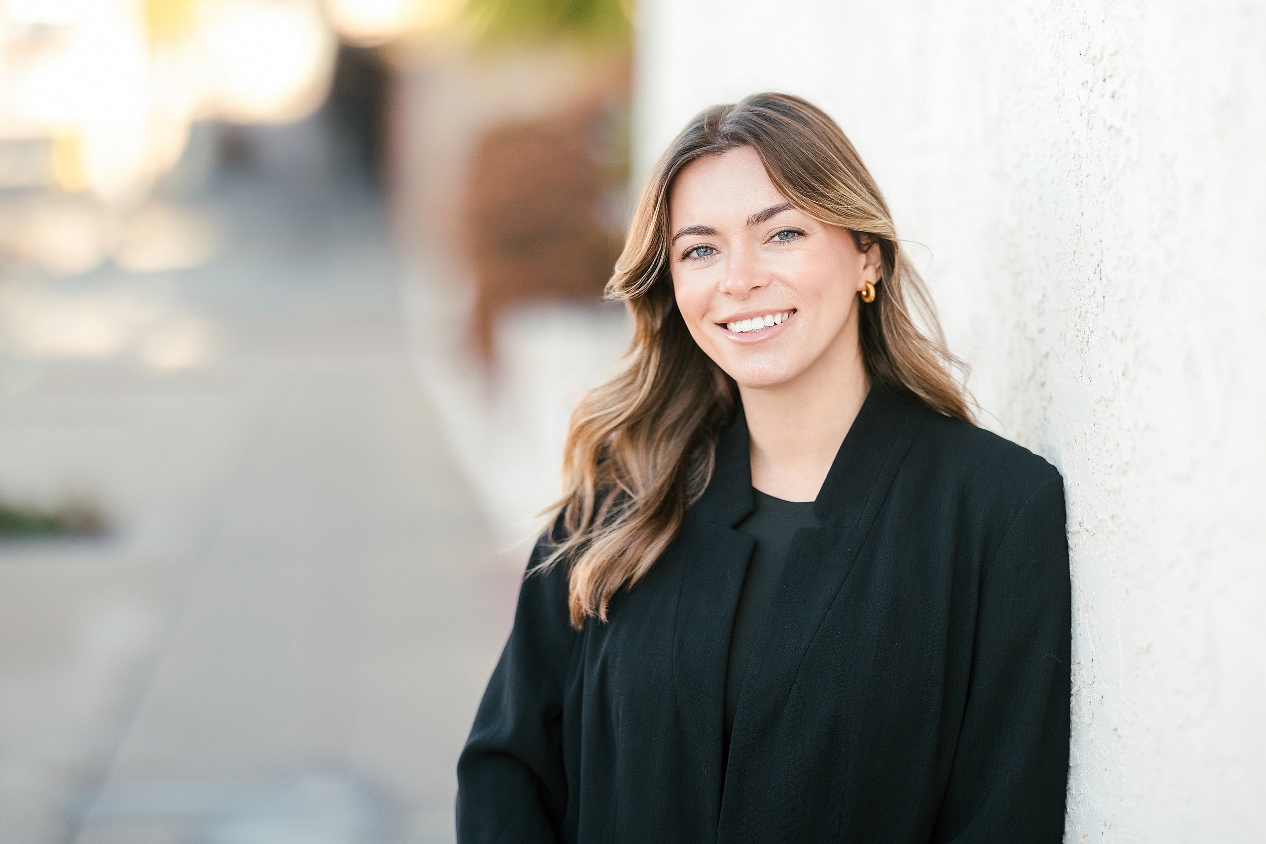 Lauren Headshots in La Mesa — 03 Lauren leans against a white wall with a blurred golden-hour city backdrop in soft focus