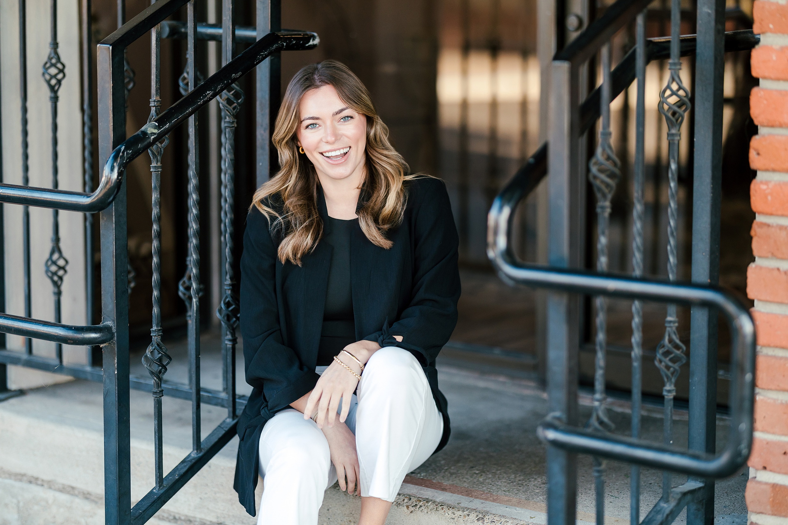 Lauren Headshots in La Mesa — 04 Woman sits on metal stairs in front of brick architecture in La Mesa, smiling