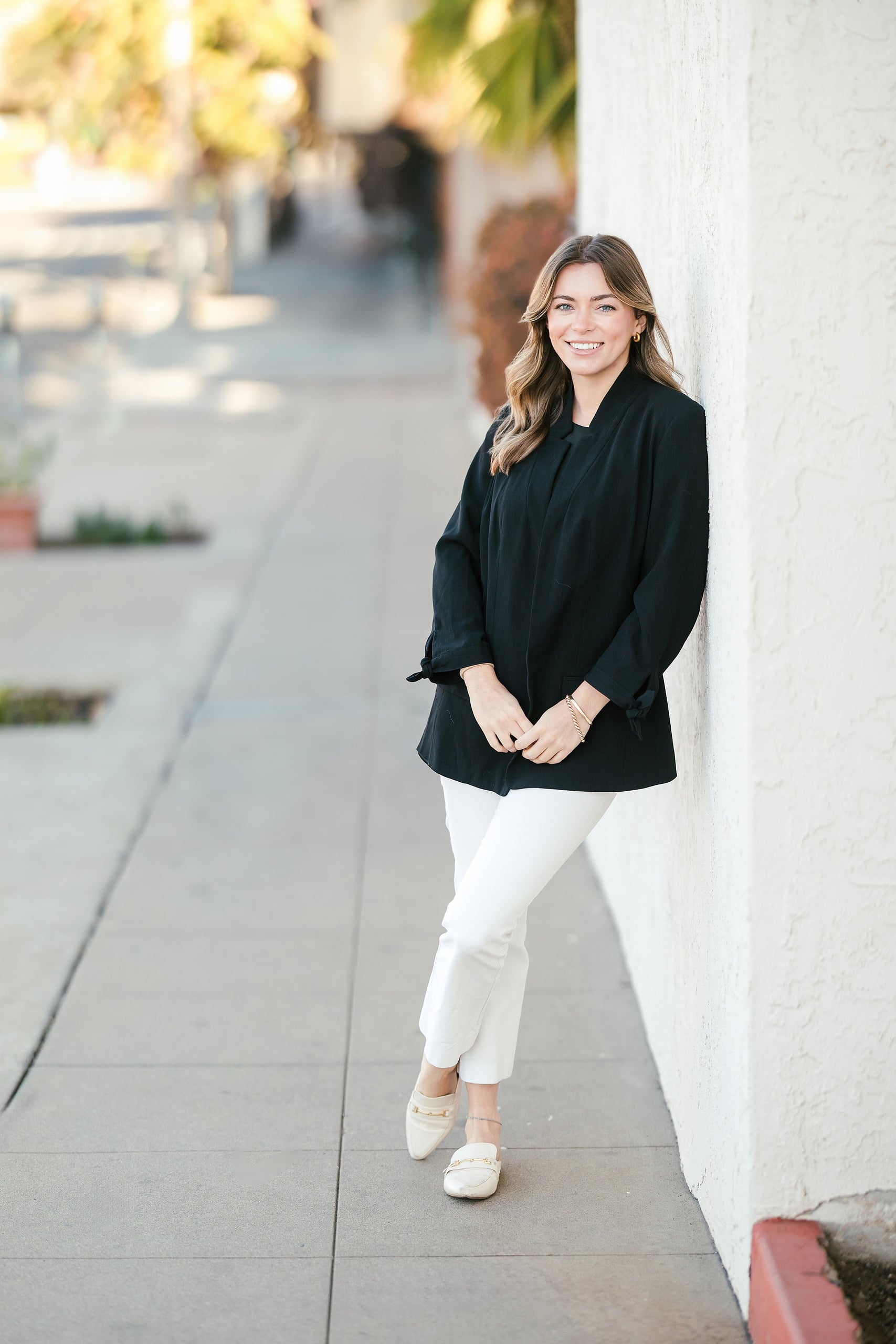 Lauren Headshots in La Mesa — 05 Professional woman stands full-length against a white wall in a black blazer and white pants