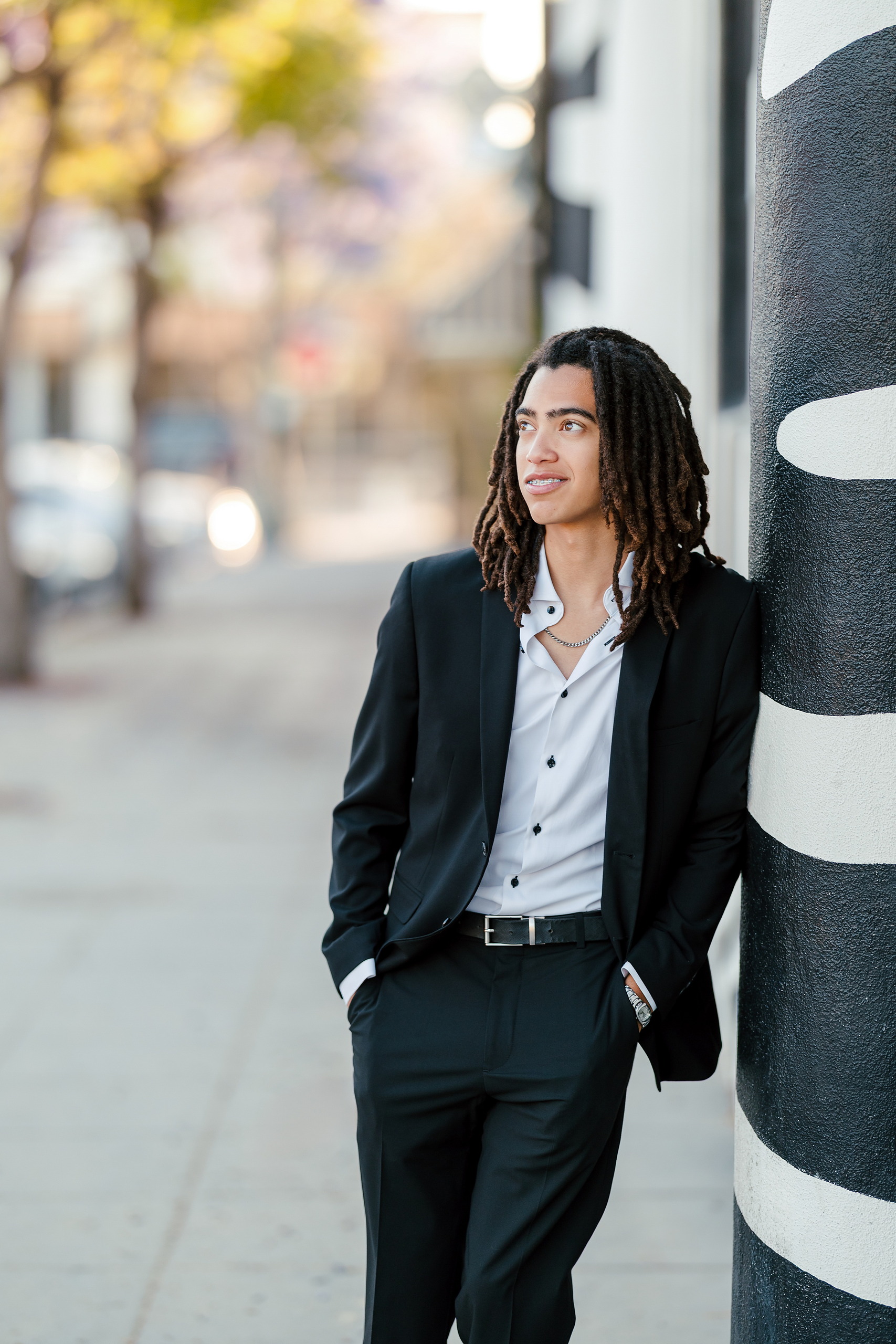Little Italy Senior Photos — 02 Joshua in black suit leaning against cylindrical post in urban setting