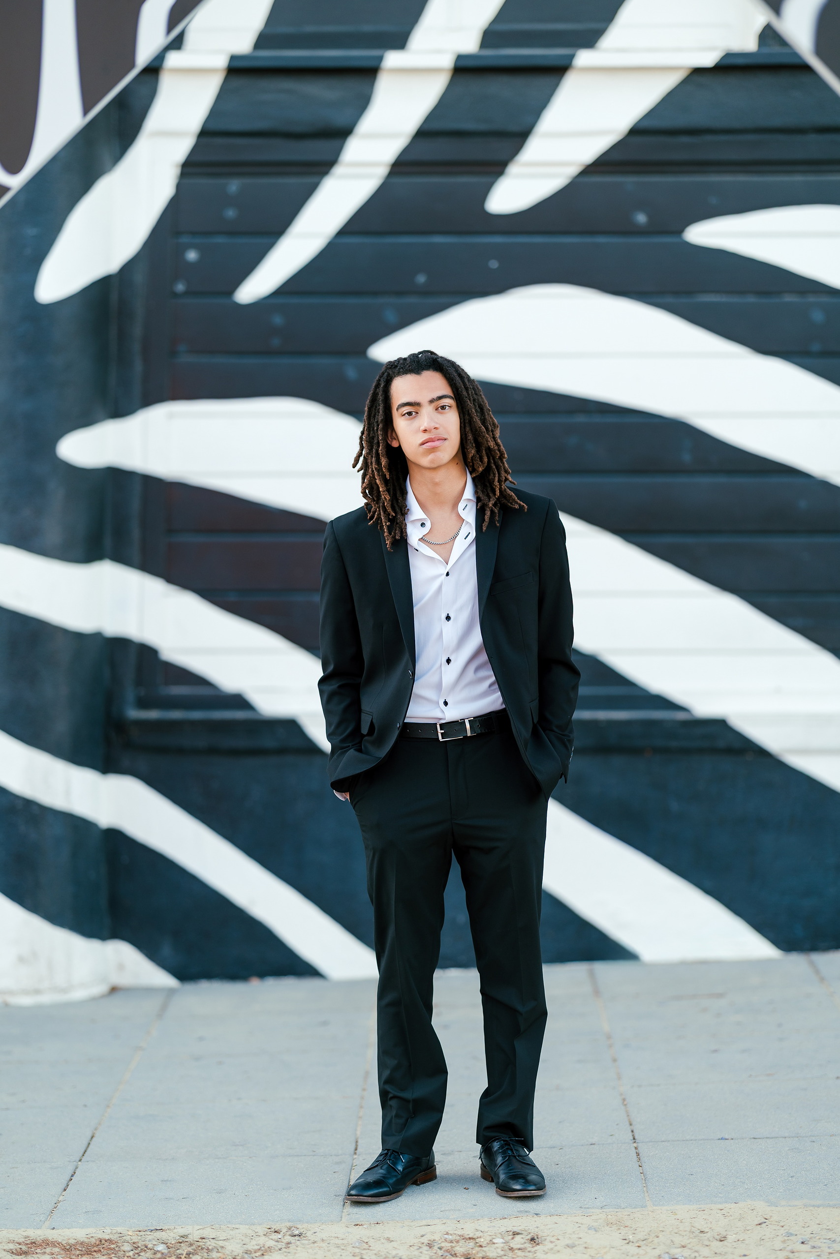 Little Italy Senior Photos — 08 Joshua in black suit against dark mural with white curved patterns