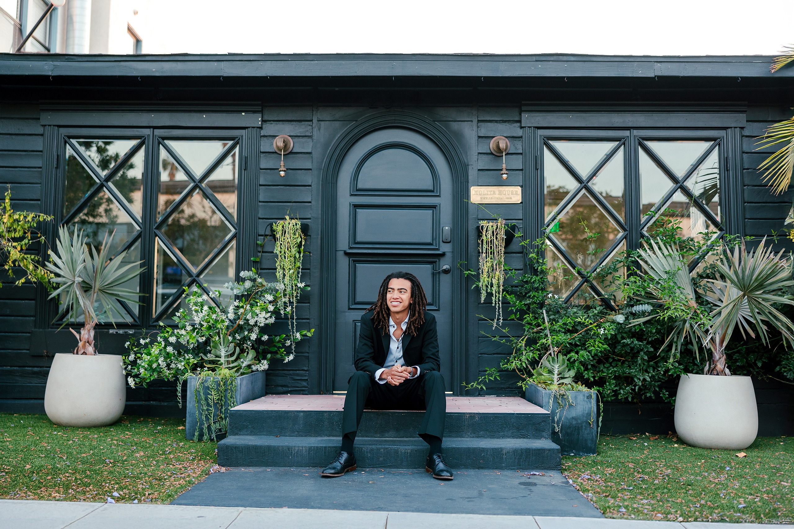 Little Italy Senior Photos — 14 Joshua seated on bench by dark building with potted plants nearby