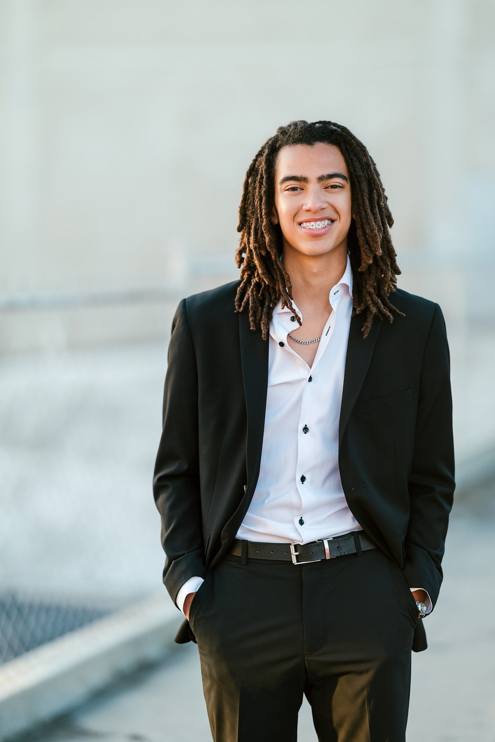 Little Italy Senior Photos — 16 Joshua grinning in black suit against cool-toned architectural wall