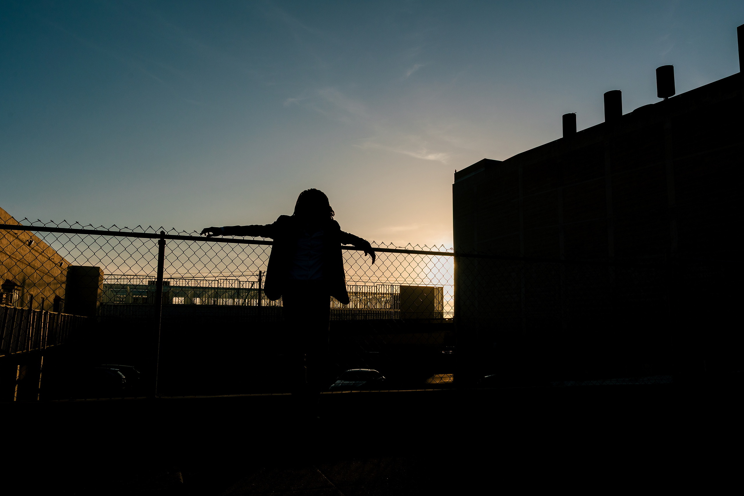 Little Italy Senior Photos — 20 Joshua silhouetted against sunset with arms on chain-link fence