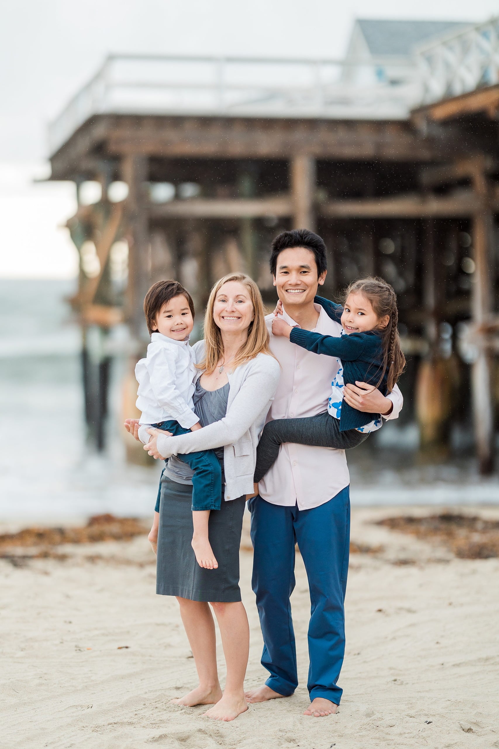 Rainy Day Beach Photo Session — 01 Woman and man hold two young children on sand at Crystal Pier Pacific Beach in San Diego