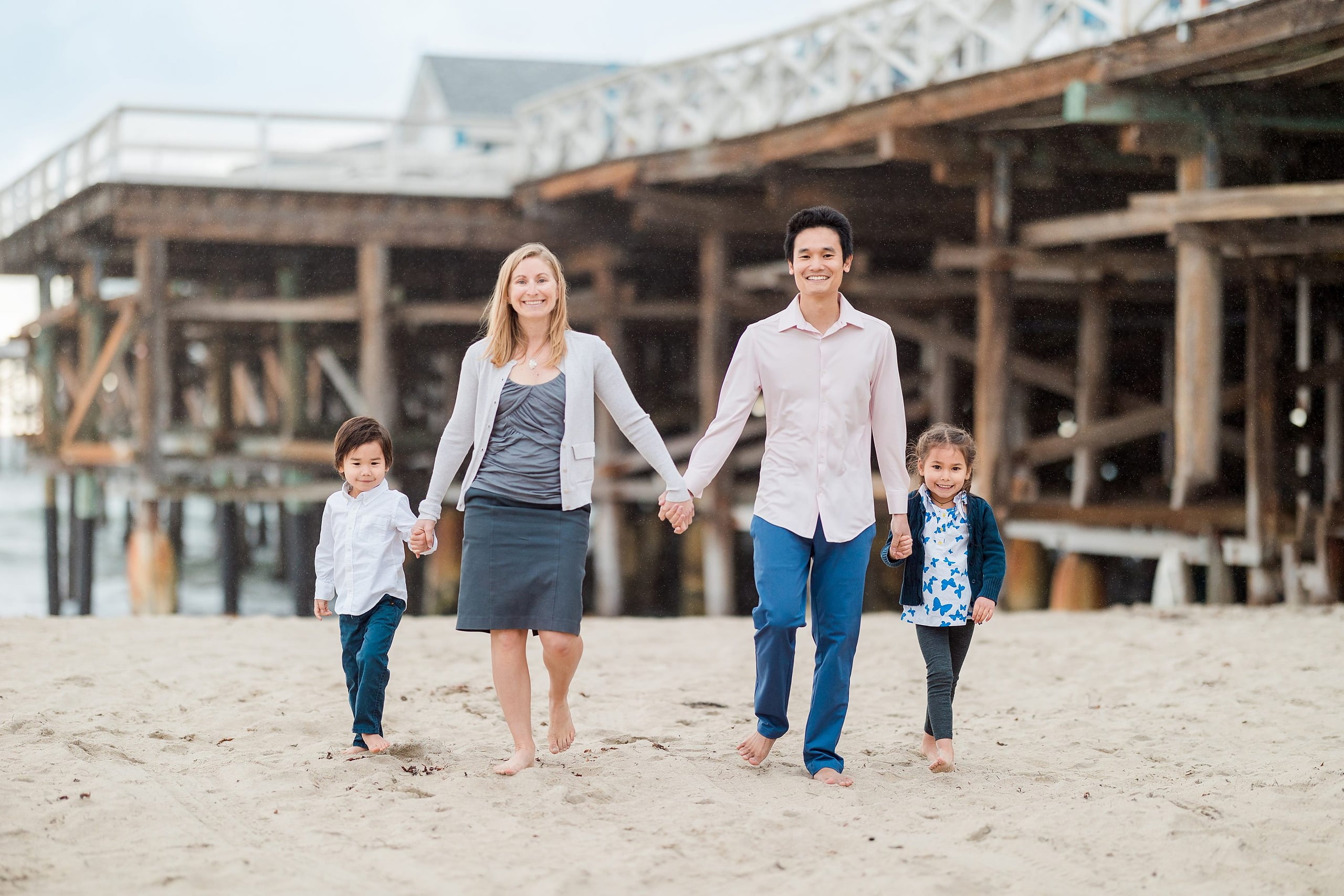 Rainy Day Beach Photo Session — 02 Family of four walk hand-in-hand barefoot across sand toward the Pacific Ocean