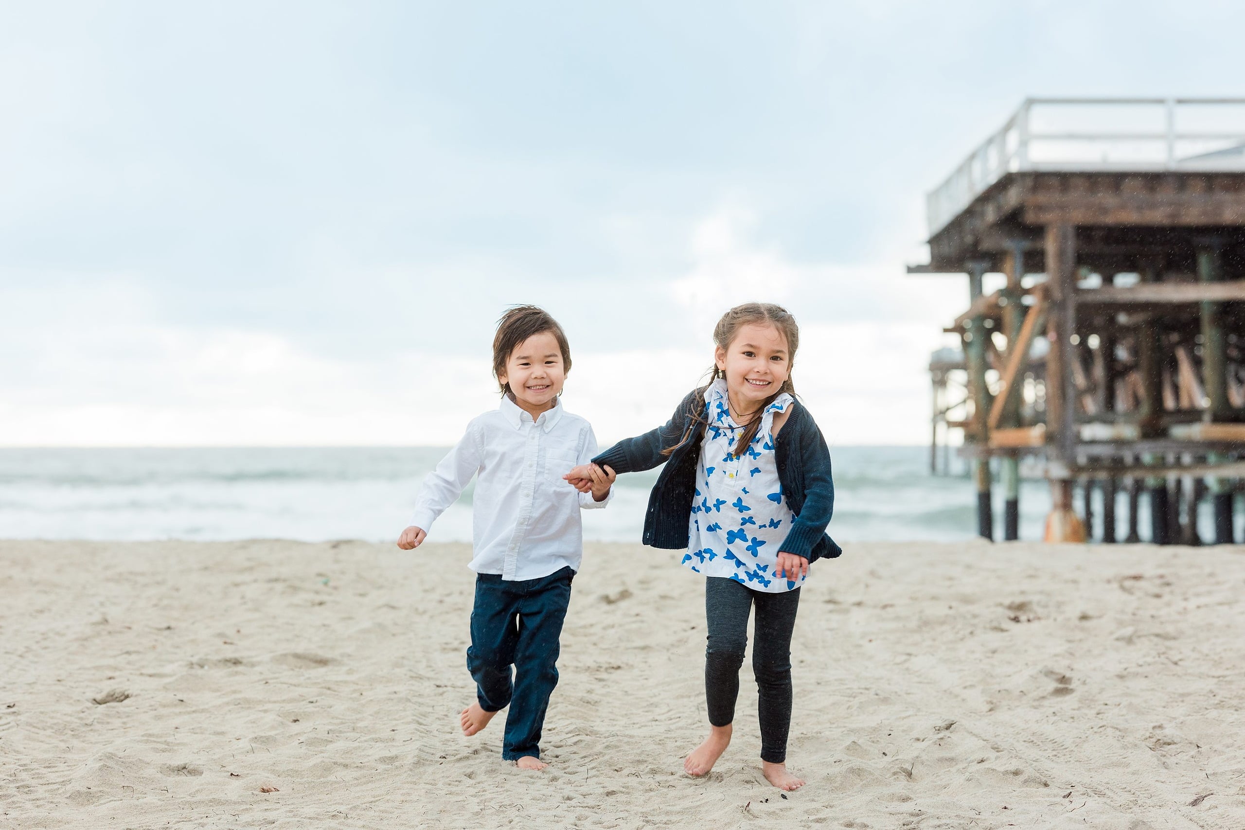 Rainy Day Beach Photo Session — 03 Two children run hand-in-hand along the beach with pier structure in background