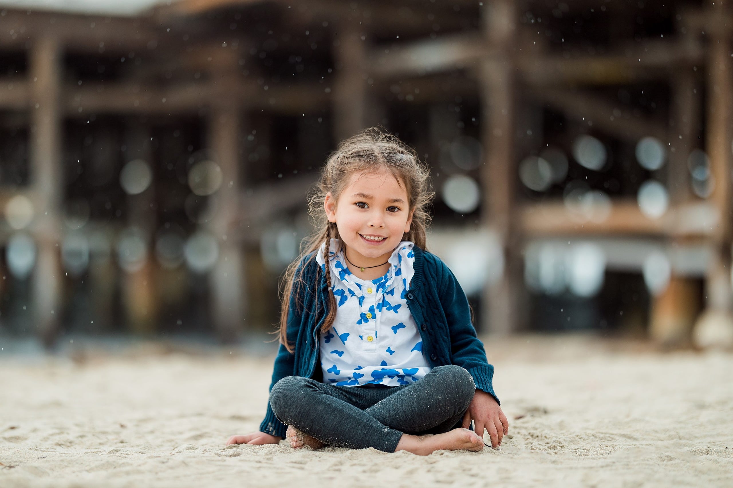 Rainy Day Beach Photo Session — 04 Two young children embrace on the sand with surf and pier visible behind them
