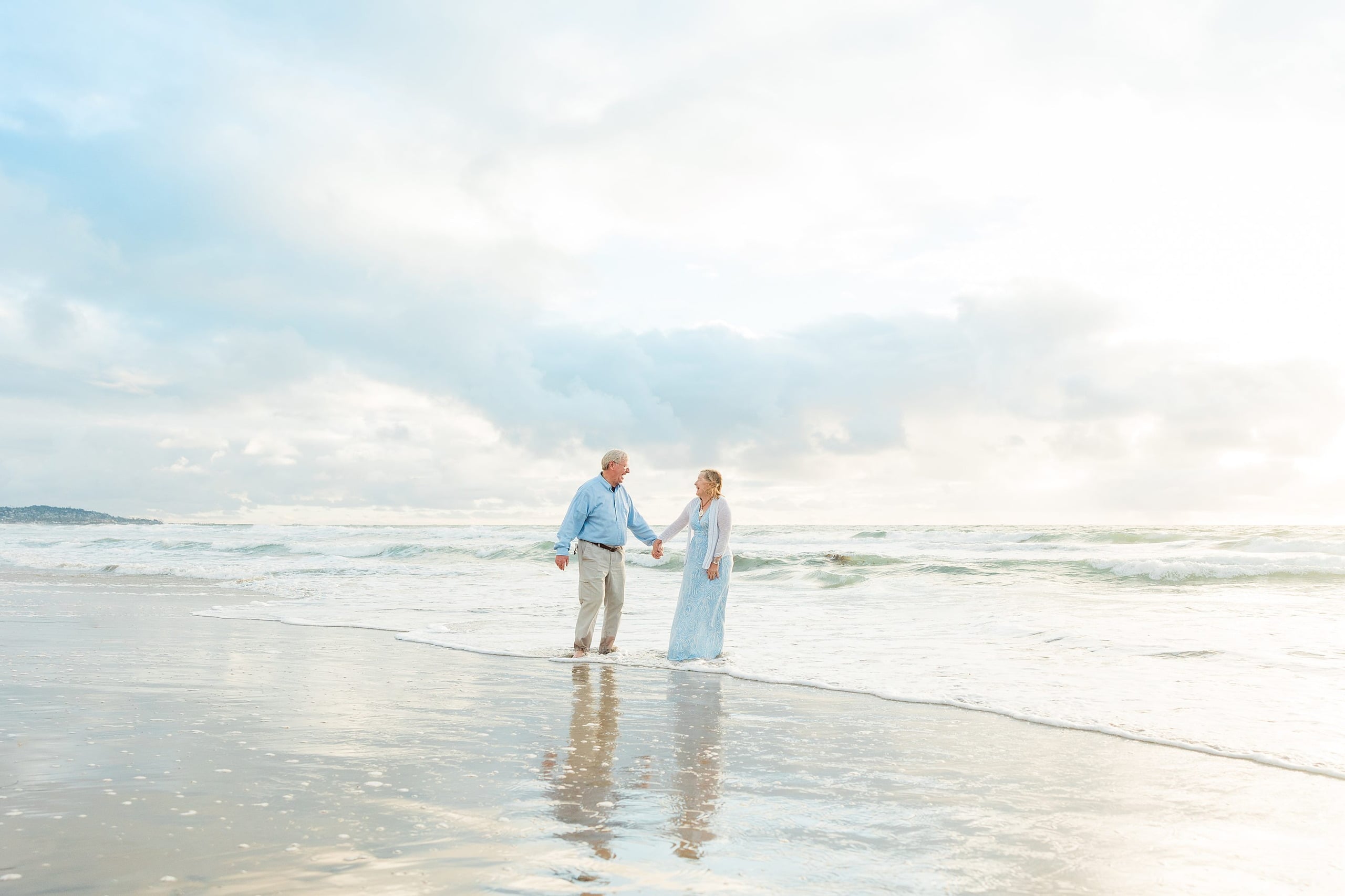 Rainy Day Beach Photo Session — 30 Couple stands holding hands wading in the surf with the ocean behind them