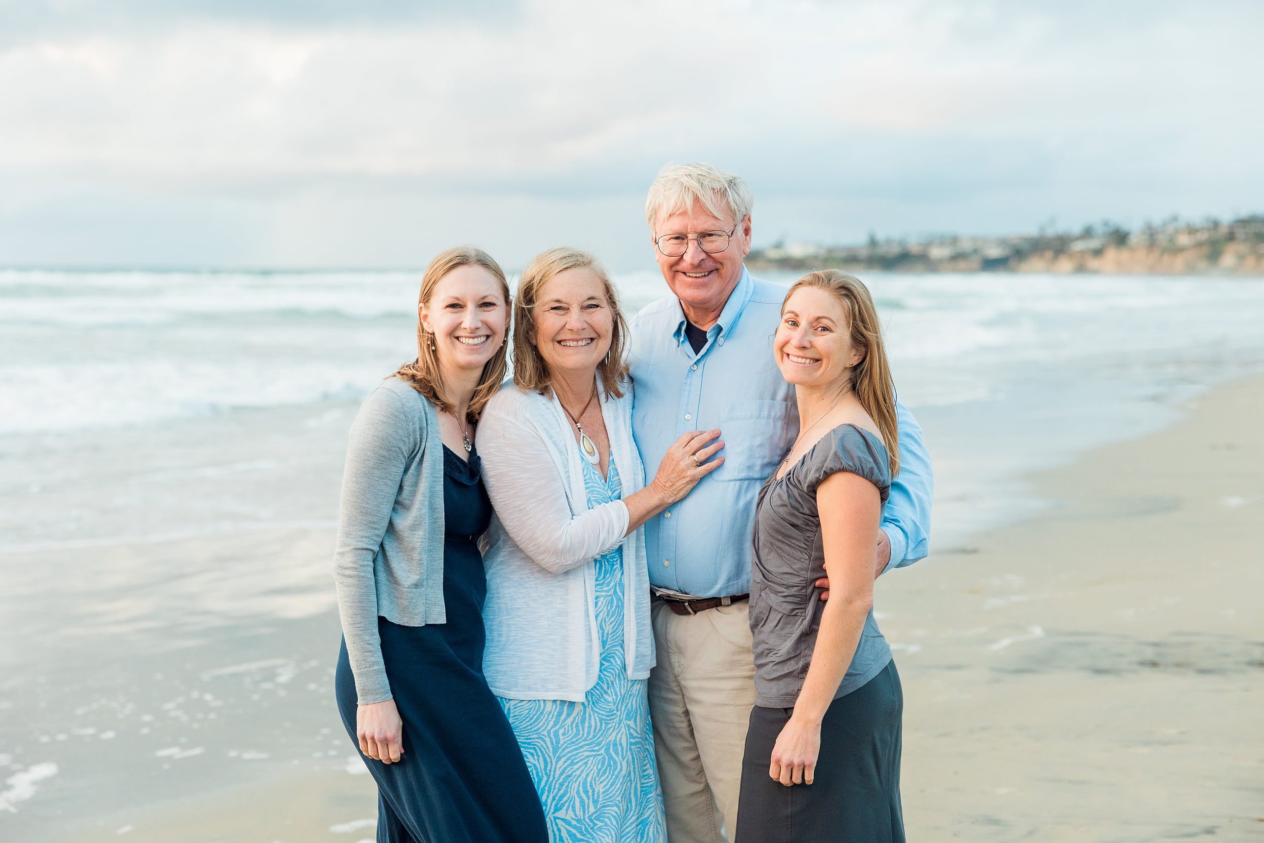 Rainy Day Beach Photo Session — 38 Two senior adults embrace while standing in shallow water facing the ocean