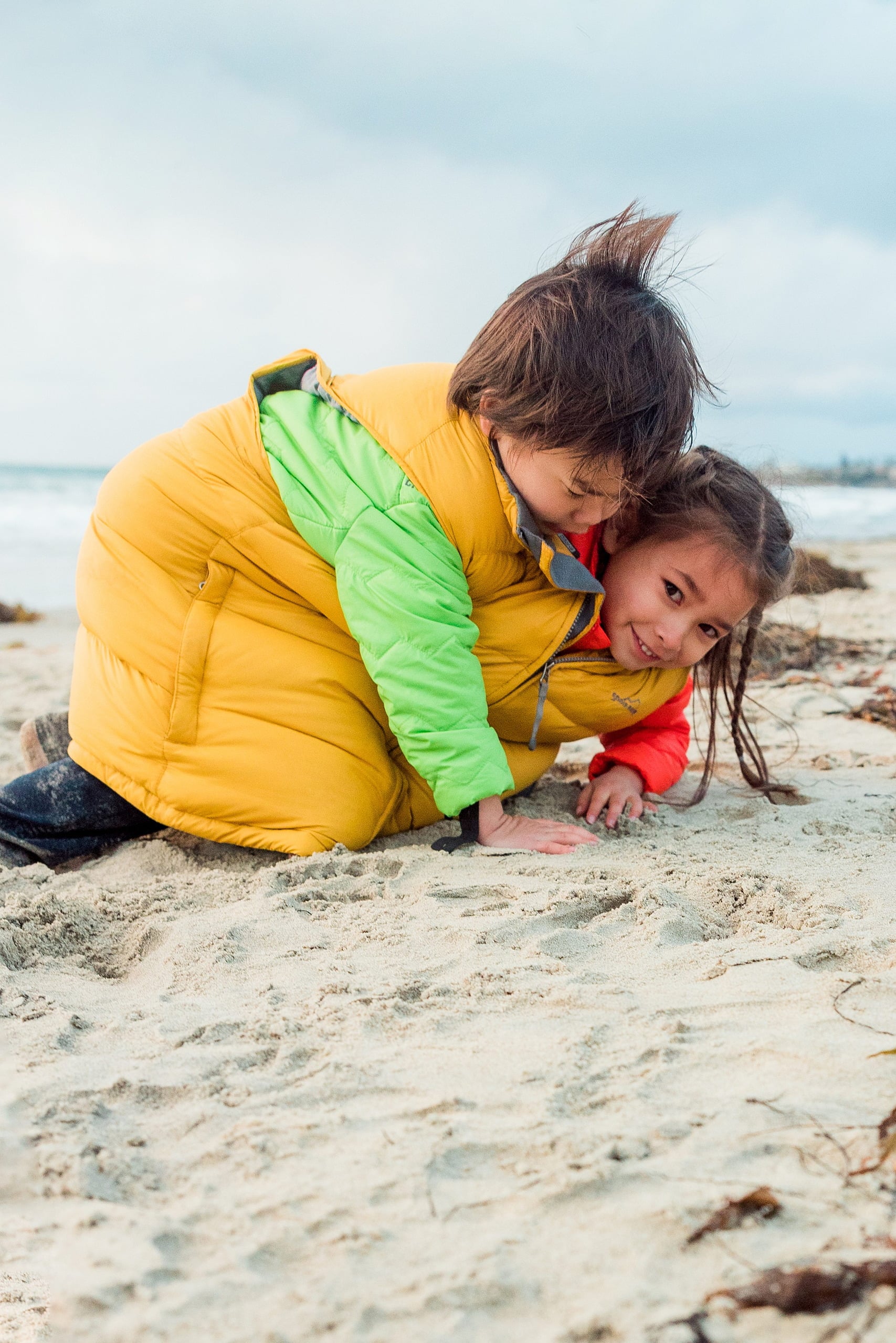 Rainy Day Beach Photo Session — 44 Two children wearing colorful puffa vests stand together on the sand at beach