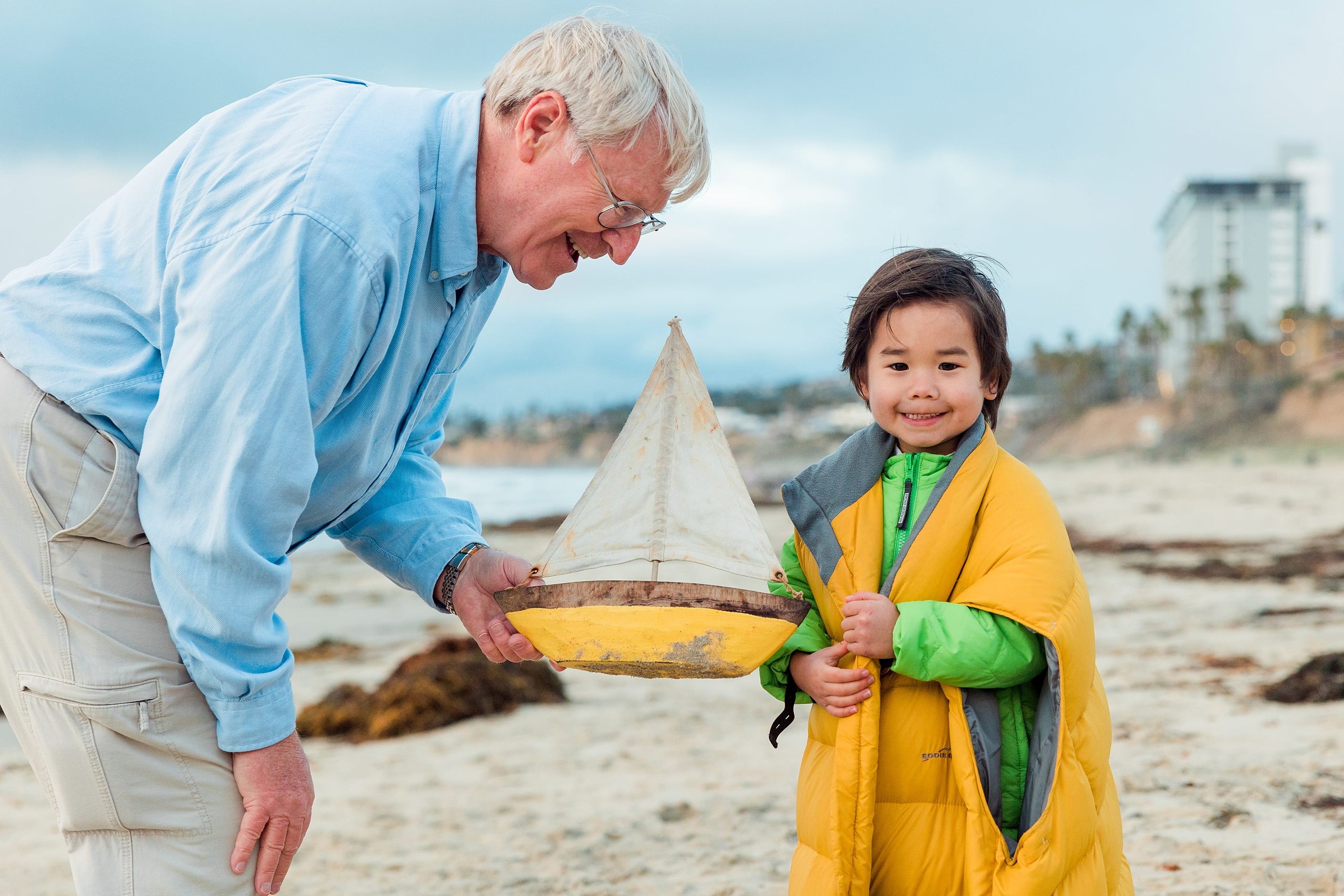 Rainy Day Beach Photo Session — 48 Child wears yellow puffa vest held by grandfather with boat toy at Pacific Beach