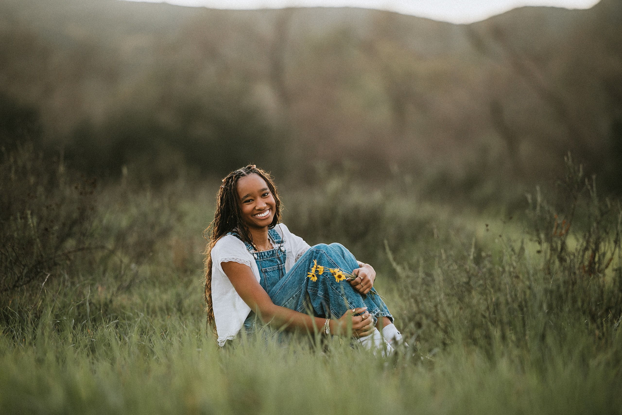 San Diego Senior Photos — 07 Kaelyn in denim overalls and white shirt kneeling in grass with yellow flowers