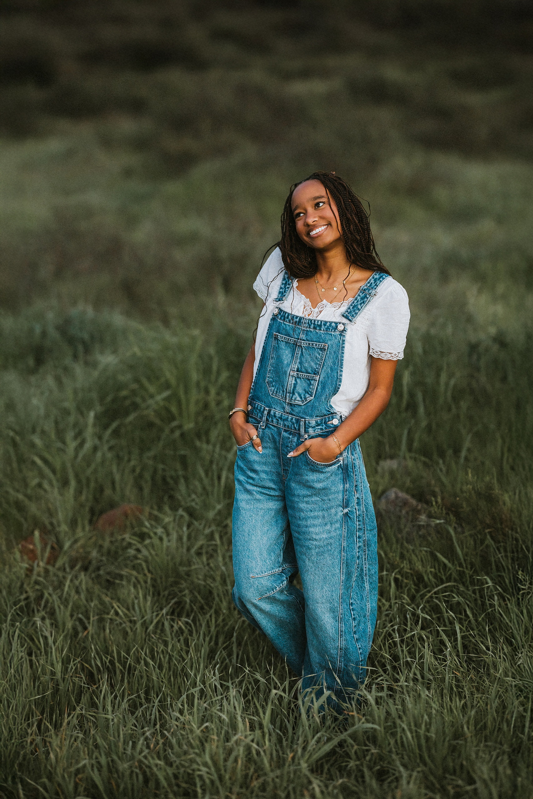 San Diego Senior Photos — 22 Kaelyn in denim overalls with white shirt, hand to chest, standing in tall grass