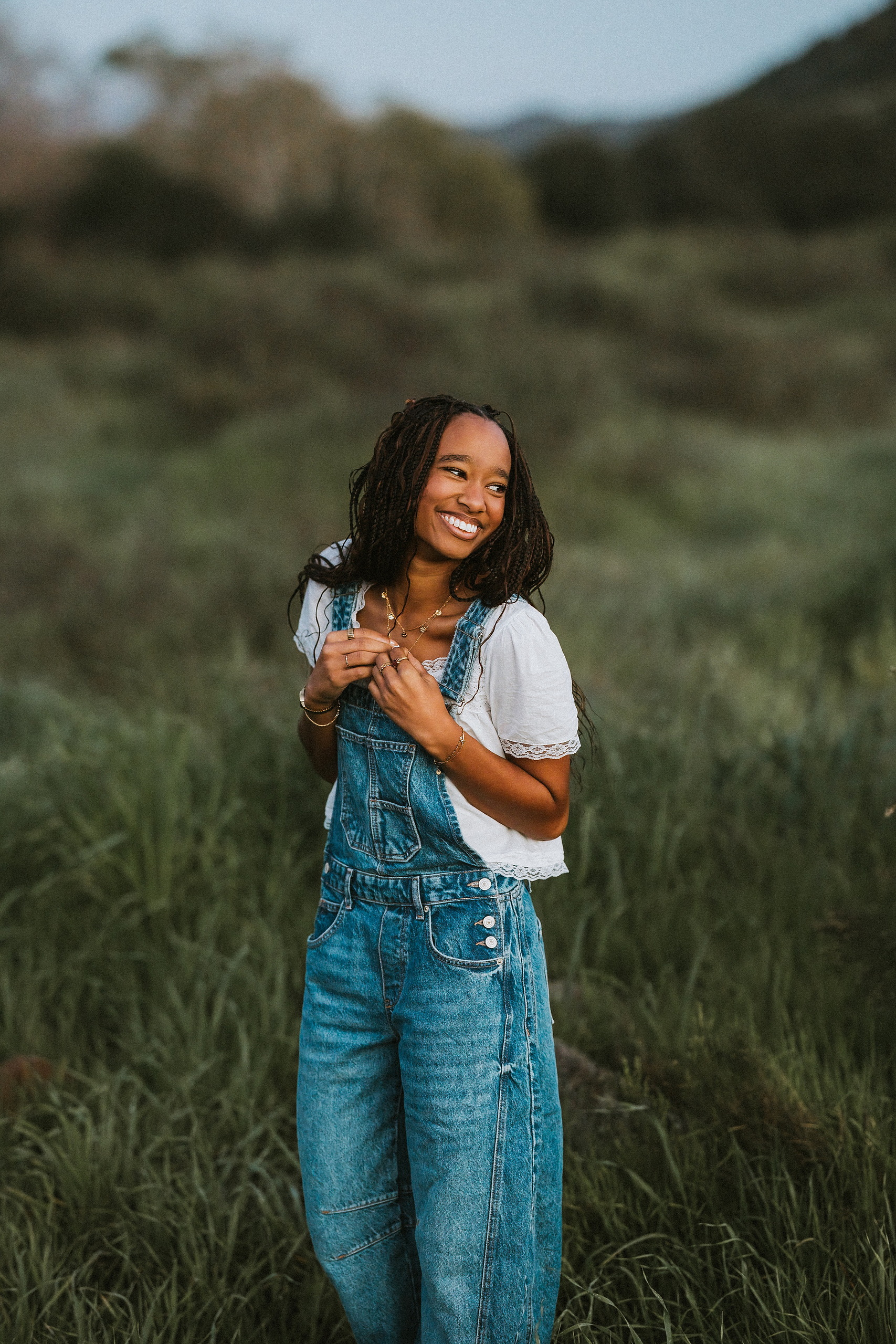 San Diego Senior Photos — 23 Kaelyn in denim overalls and white shirt with vest, standing in grassy field