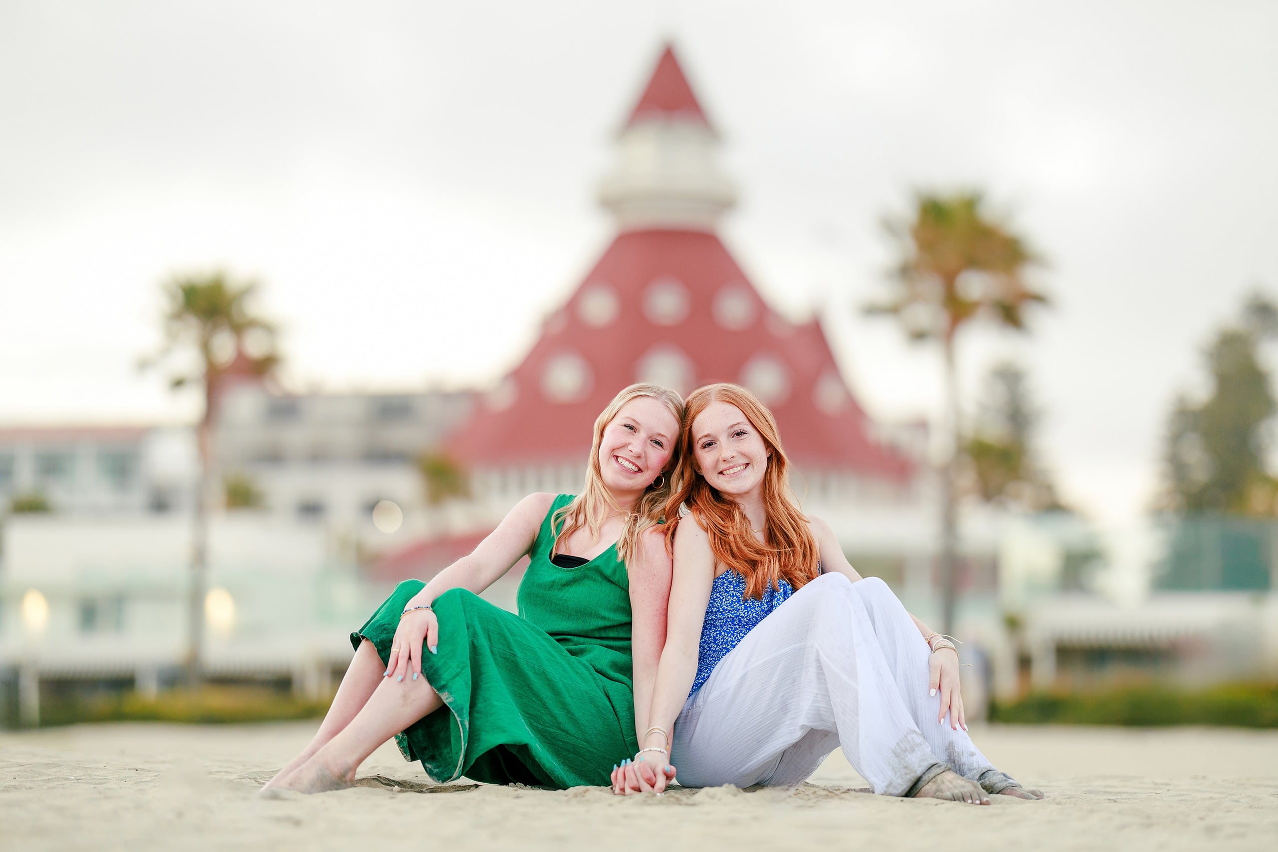 Sisters & Senior Portraits at the Del — 41 Two sisters sitting together on beach sand with historic hotel building behind