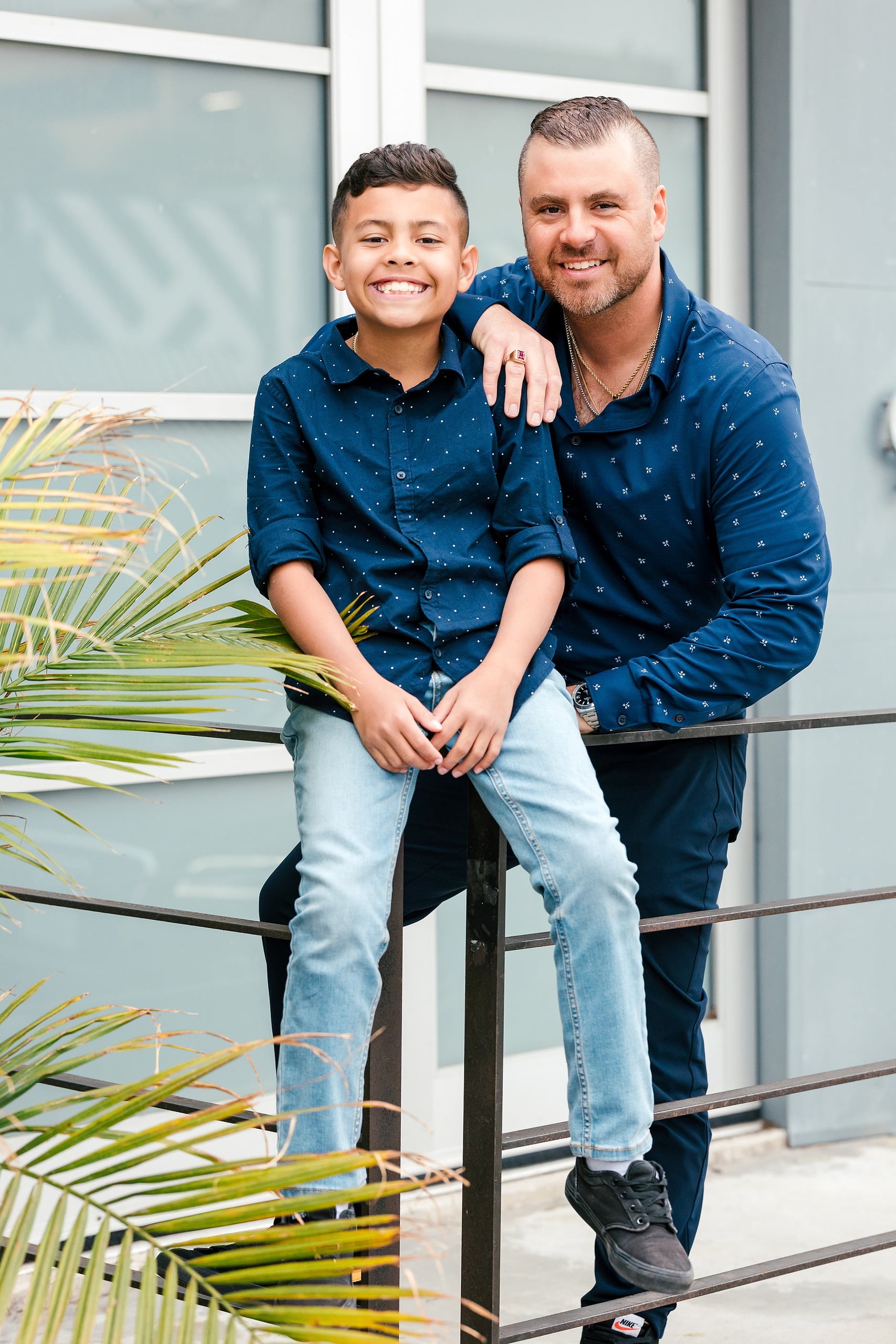 Spring Break Surprise: Why This Mom Booked a Secret Photo Session — 10 Child and man sit on metal railing with palm plant, modern building behind