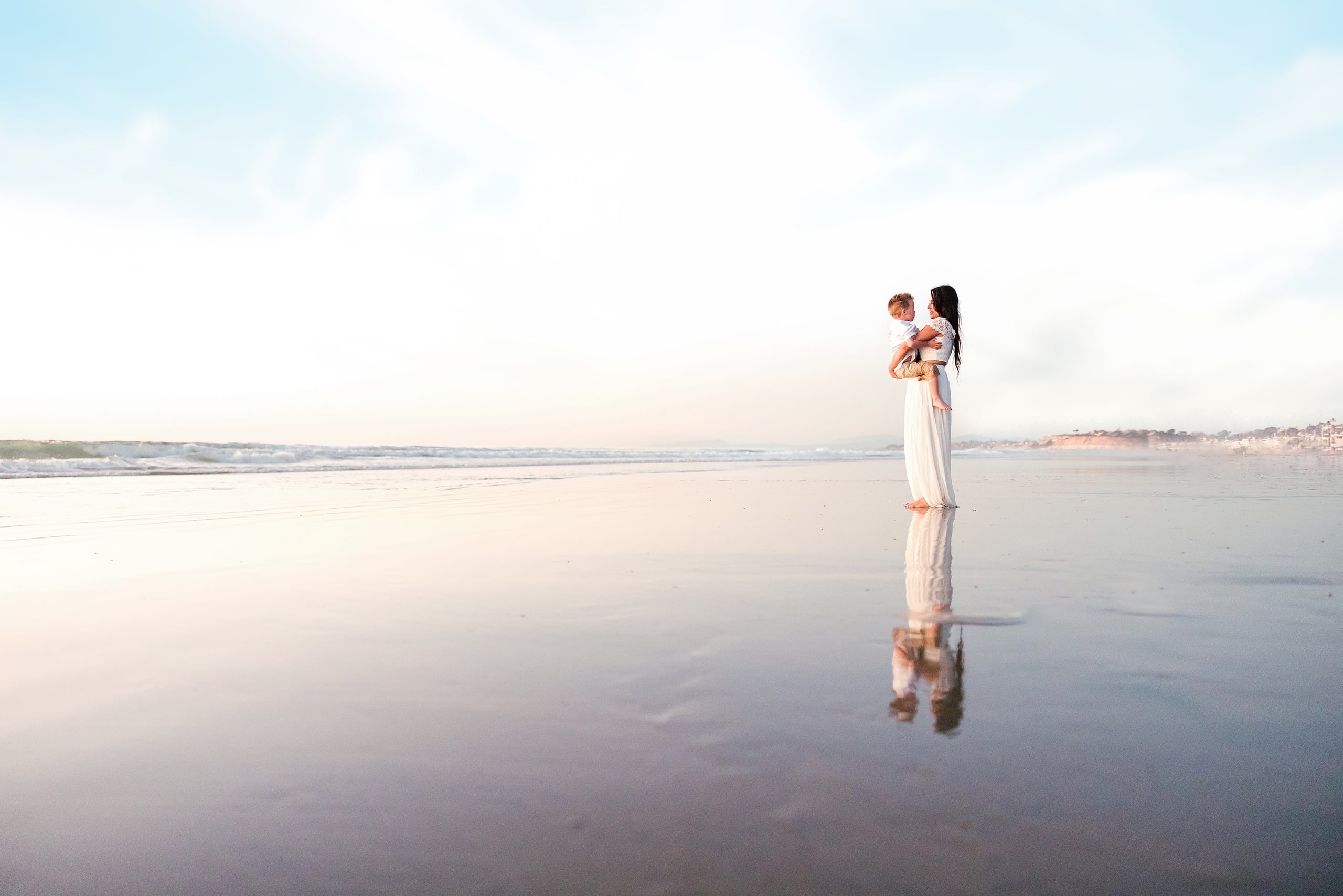 The Day the Sun Stood Still — 17 Woman holding toddler in white dress on wet beach sand at Del Mar sunset