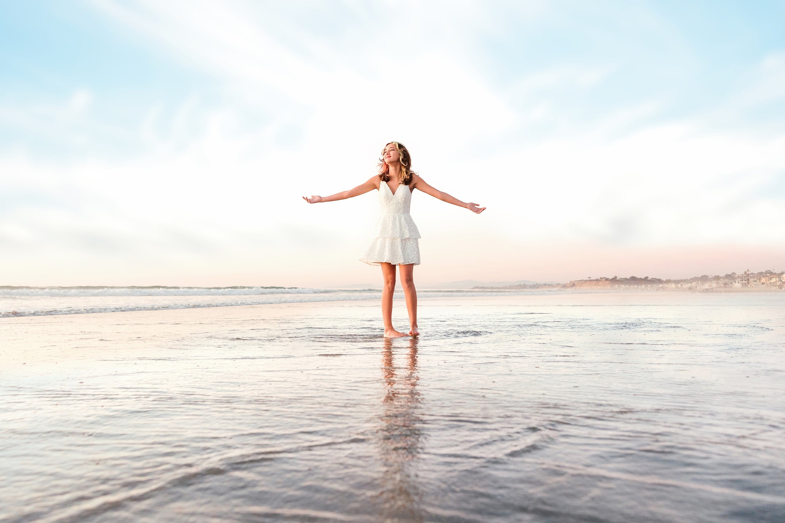 The Day the Sun Stood Still — 20 Teen in white dress with arms outstretched on wet beach sand at sunset