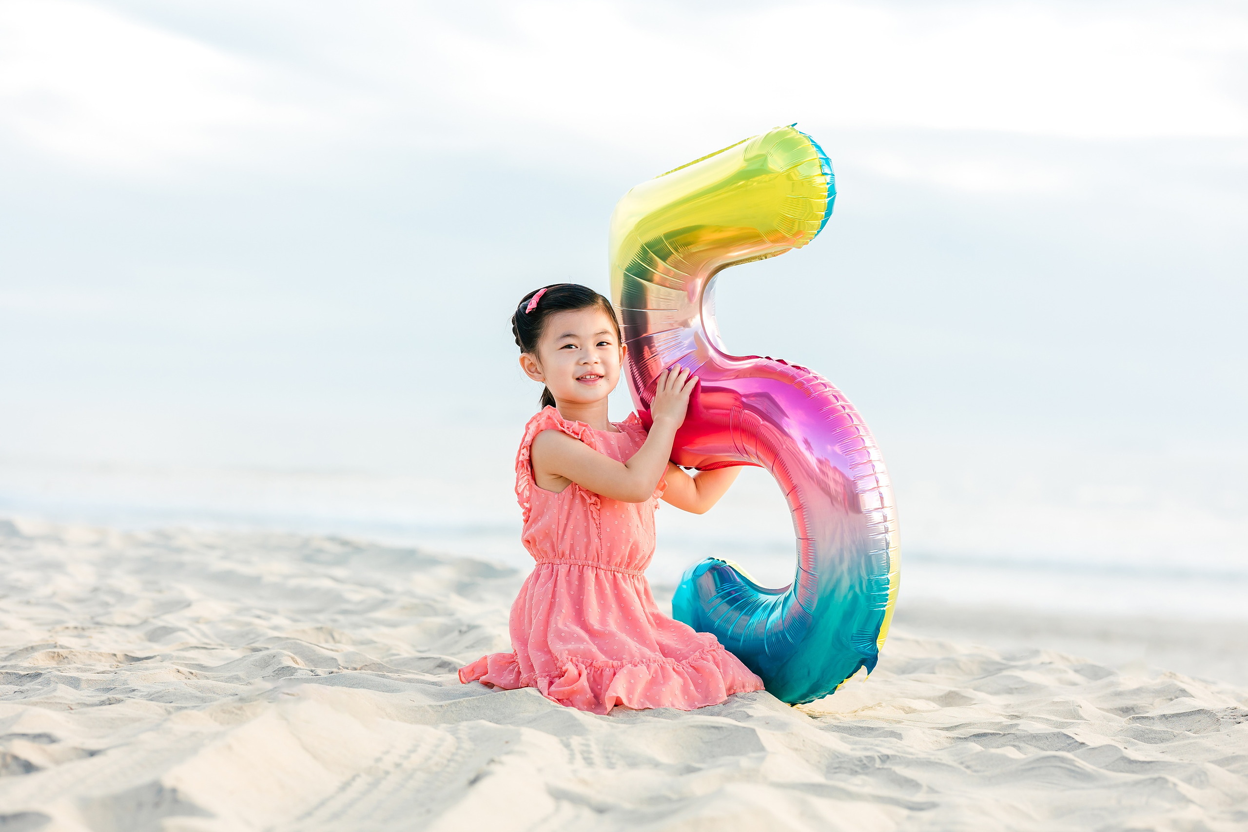 Turning Five on Coronado — 01 Ava kneeling on sand holding a colorful fifth birthday balloon at beach