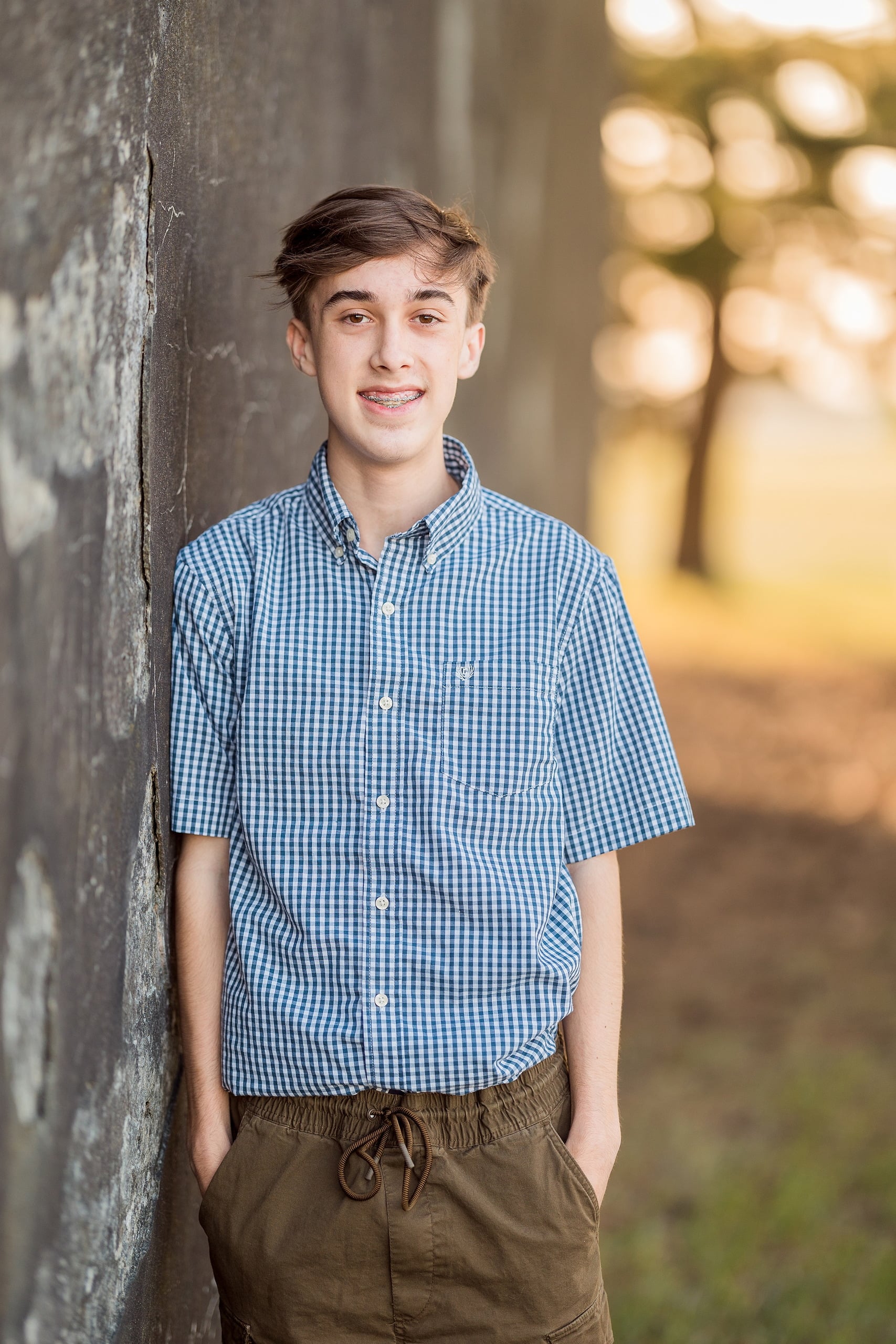 Zach’s Senior Portraits — 02 Zach standing against aged wall in blue checkered shirt, golden light