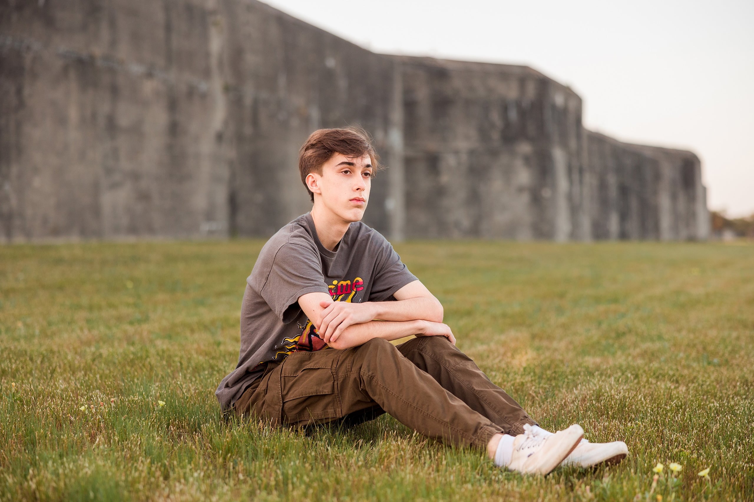 Zach’s Senior Portraits — 18 Zach sitting in grass field with Fort Monroe Battery behind him