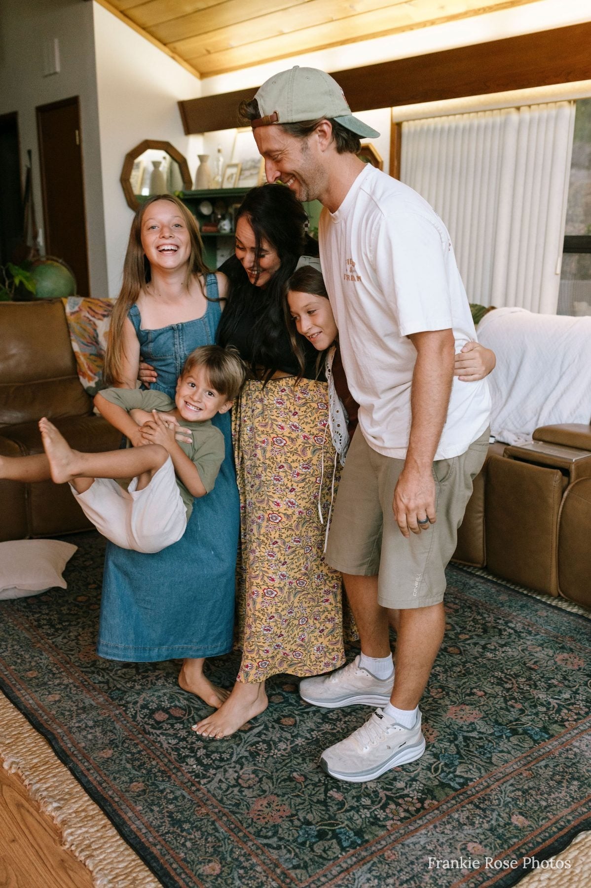 Family of five laughs together on rug, dad holds child upside down