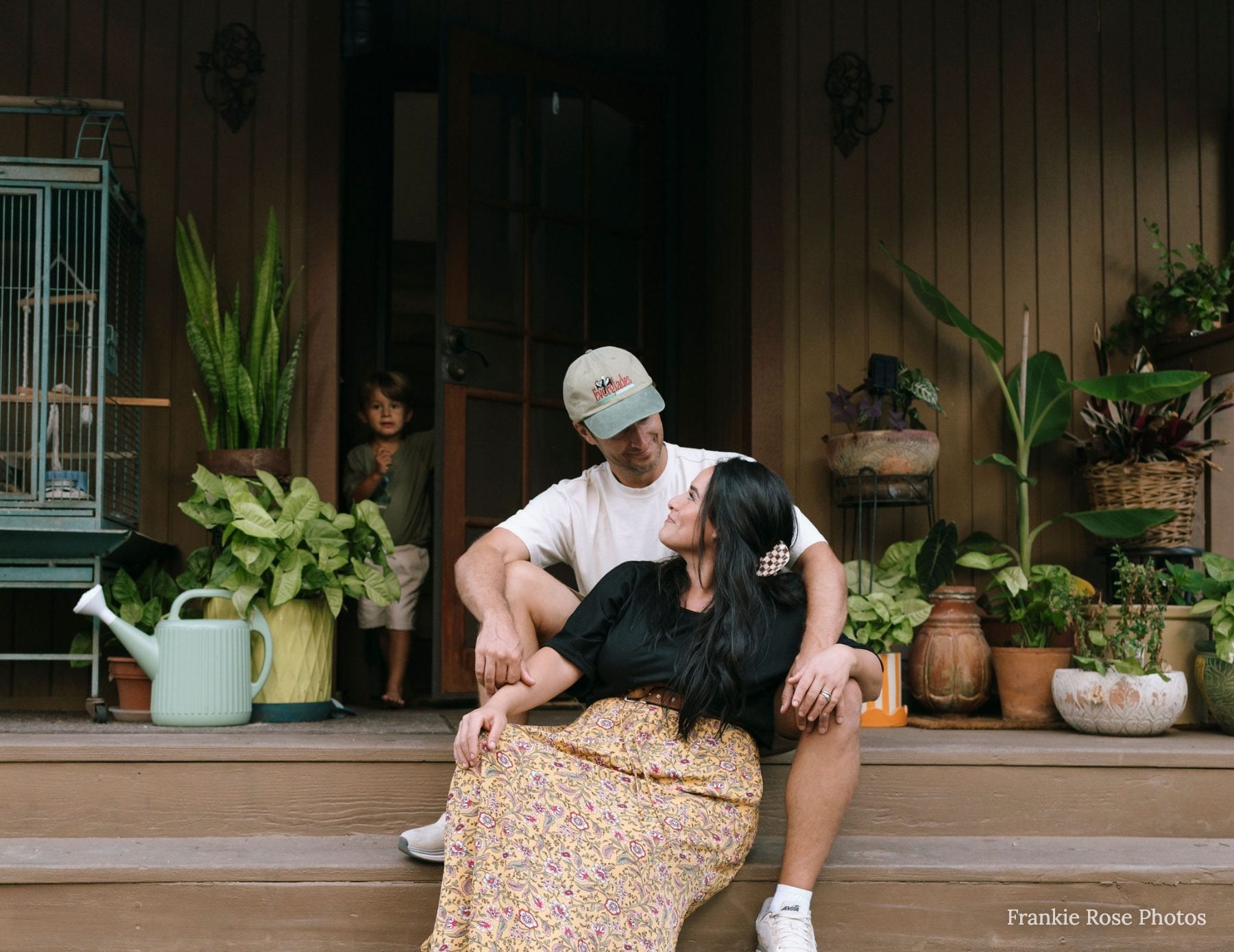 Couple sits close on porch steps among potted plants, son peeking behind