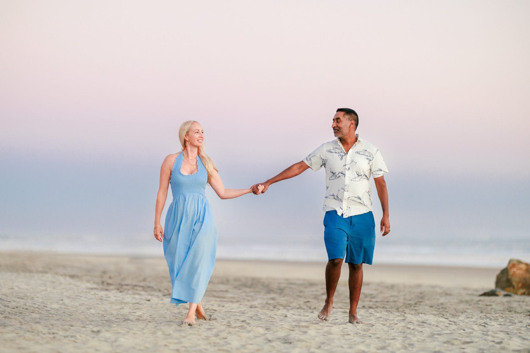 Couple holds hands walking barefoot along Coronado beach at dusk