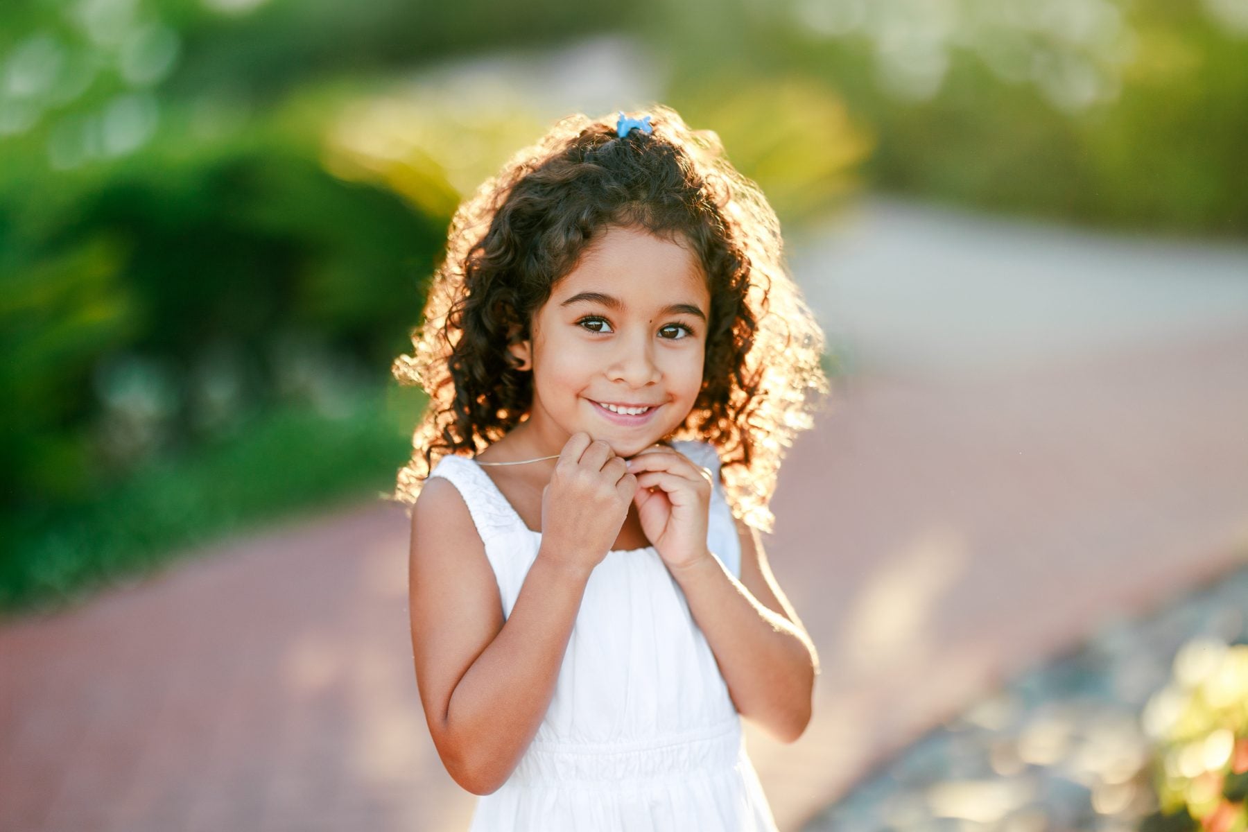 Curly-haired girl in white dress clasps hands, smiling for San Diego photographer