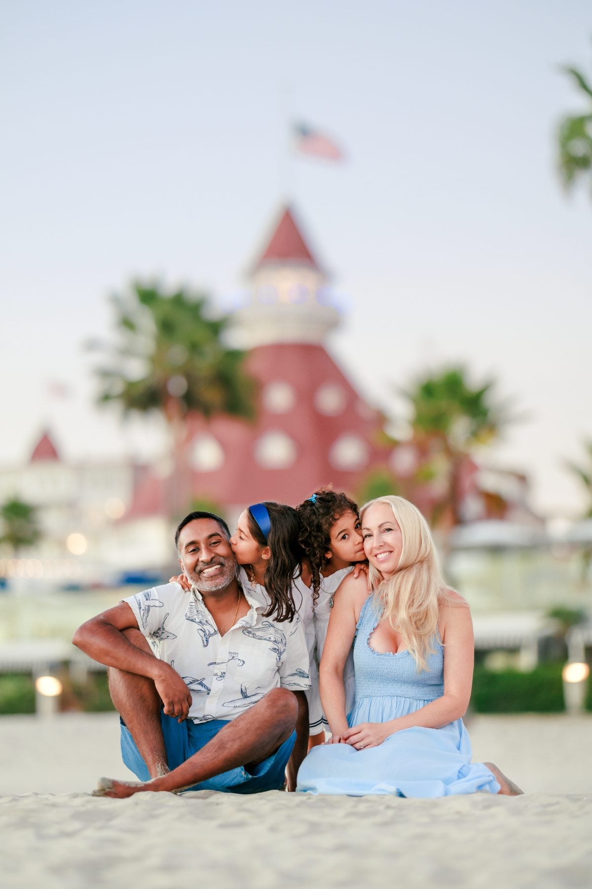 Family of four sits on sand as daughter kisses dad's cheek, Hotel del Coronado behind