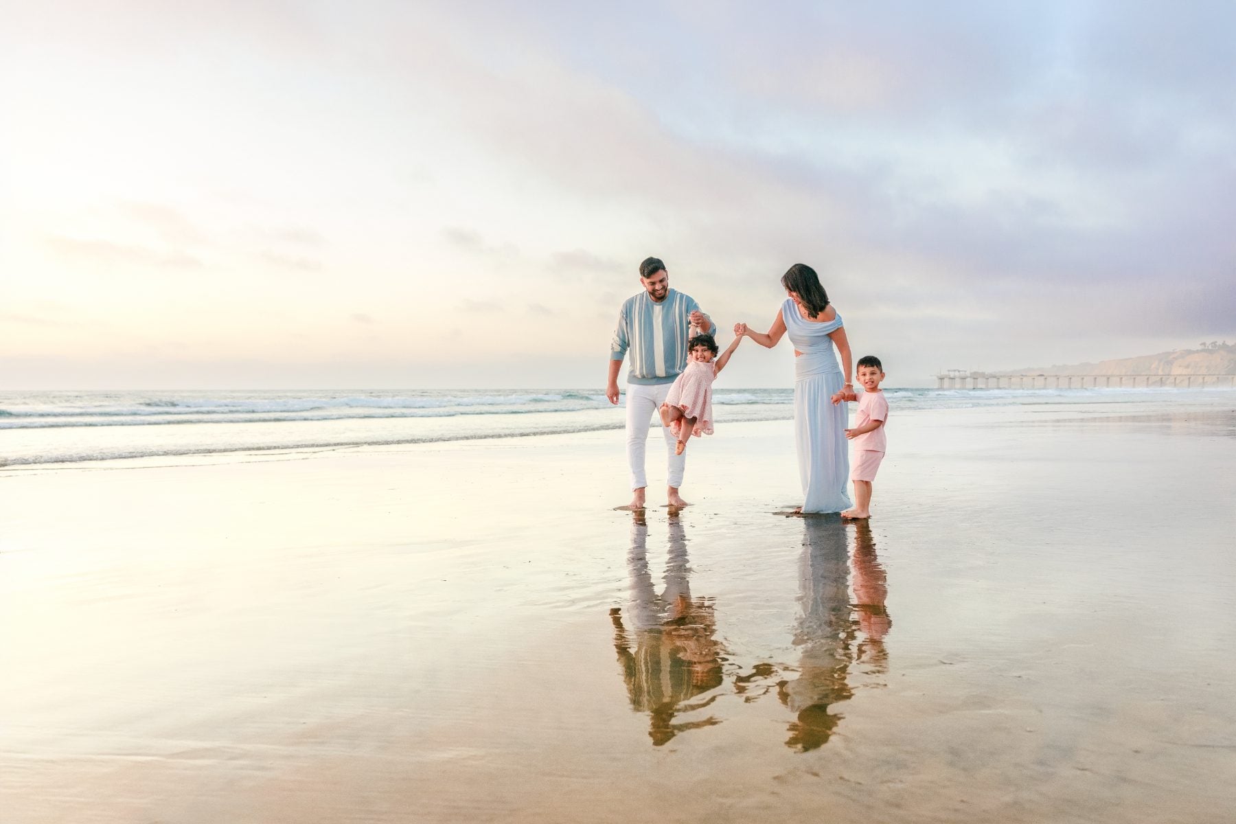 Parents and twins hold hands walking on wet sand near La Jolla pier