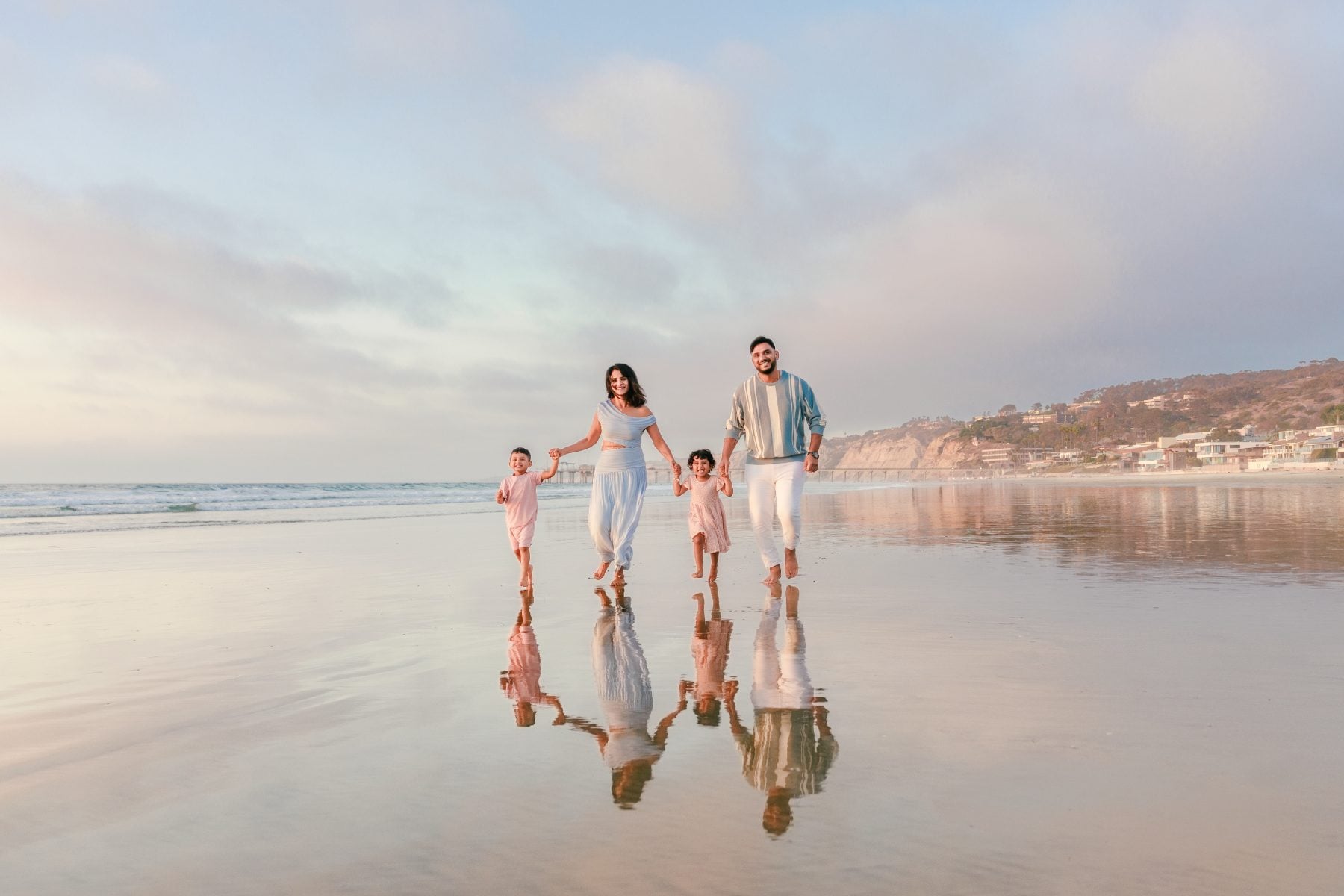 Family of four strolls along shoreline, reflections visible in wet sand, La Jolla