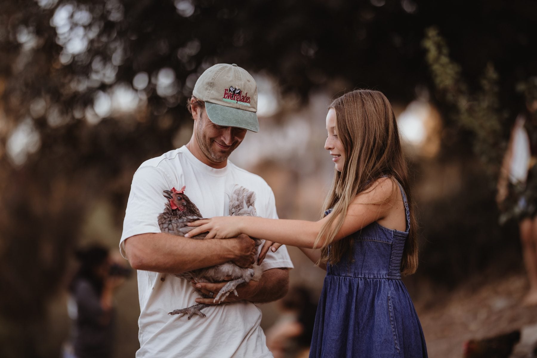 Father in cap holds chicken while daughter reaches out at their Lakeside home