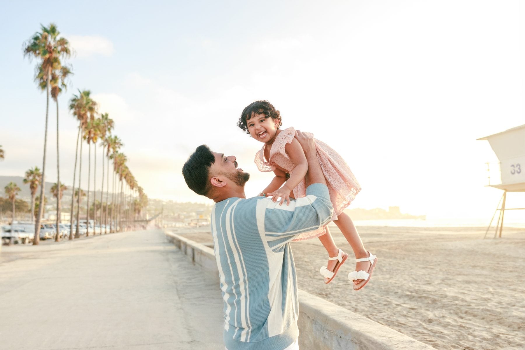 Father lifts giggling toddler girl near palm trees and lifeguard tower