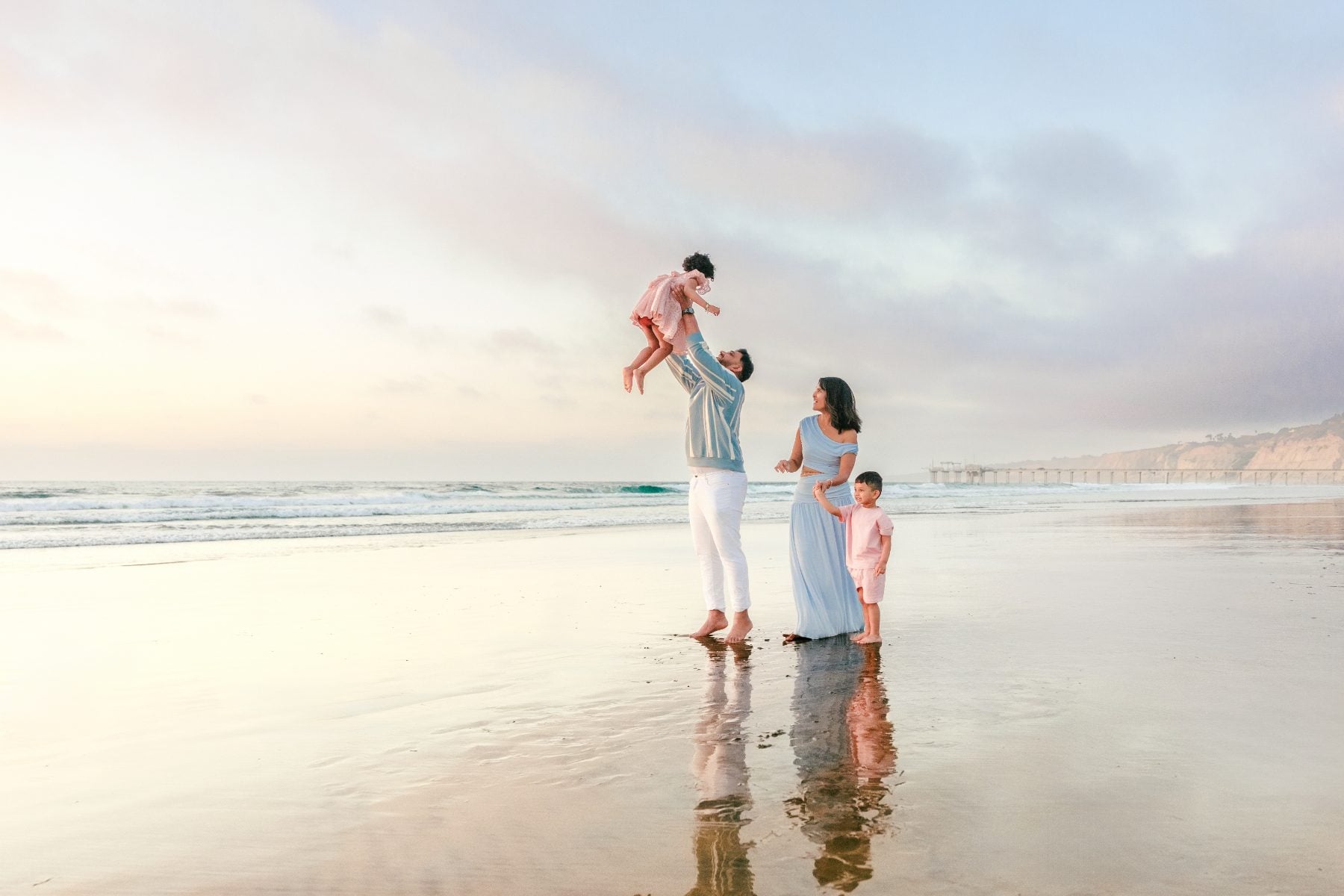 Dad tosses toddler into the air as mom and son watch, La Jolla pier behind