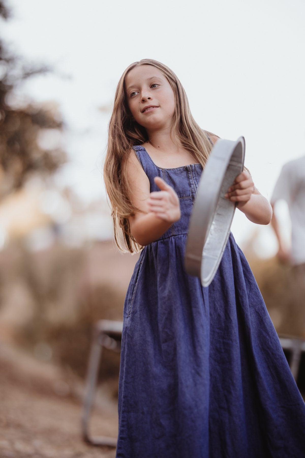 Girl in blue dress swings a metal pan while playing outside
