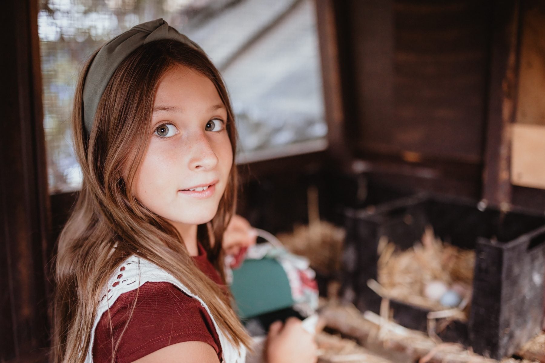 Young girl in headband glances back inside a rustic chicken coop