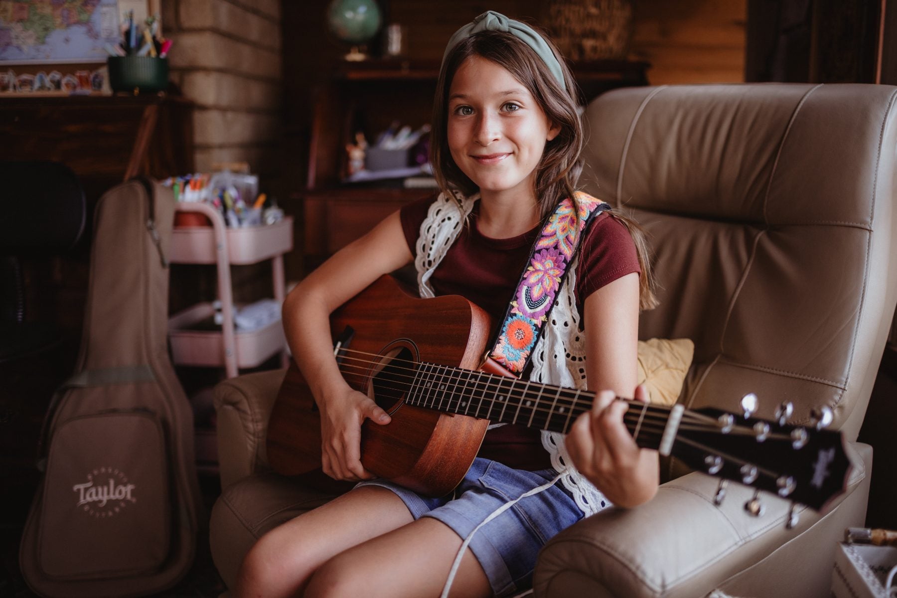 Young girl smiling while playing an acoustic guitar in a cozy living room chair