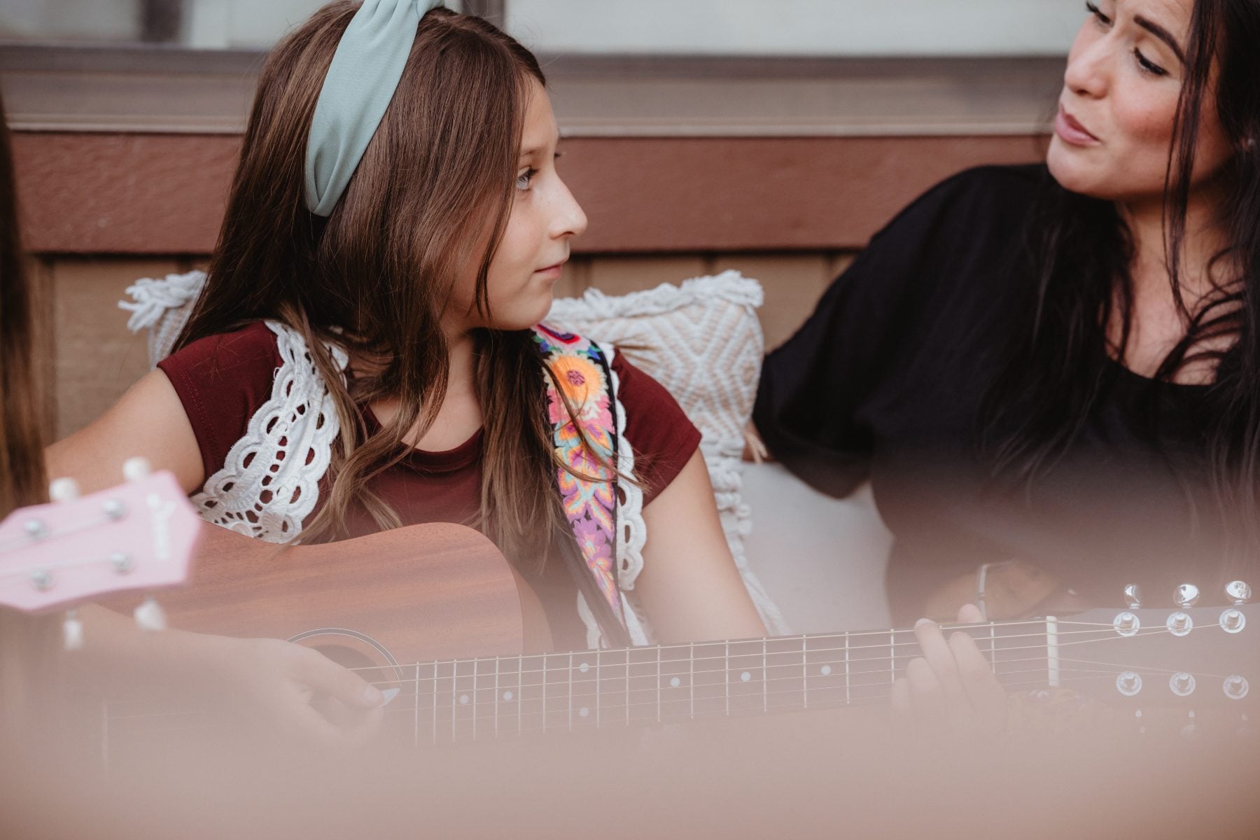 Girl plays acoustic guitar while woman listens on porch cushions