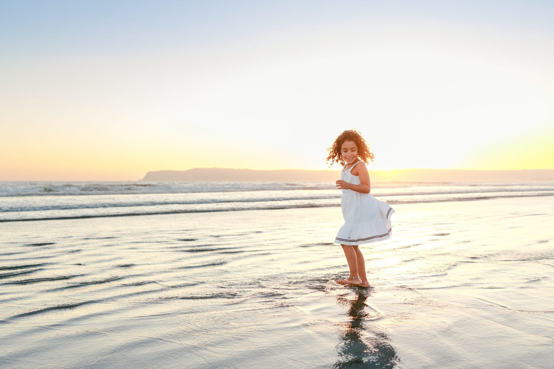 Young girl twirls in a white dress along the shoreline at dusk