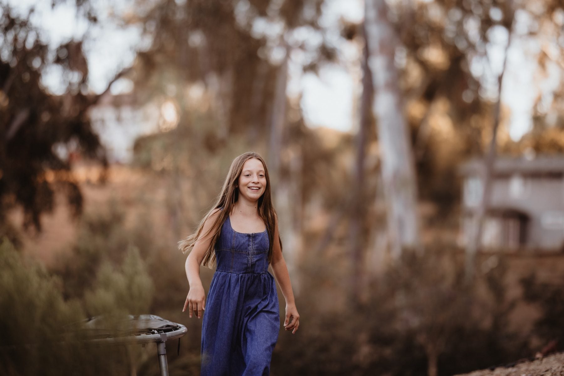 Girl in blue jumpsuit walks smiling through sunlit trees, San Diego family photographer