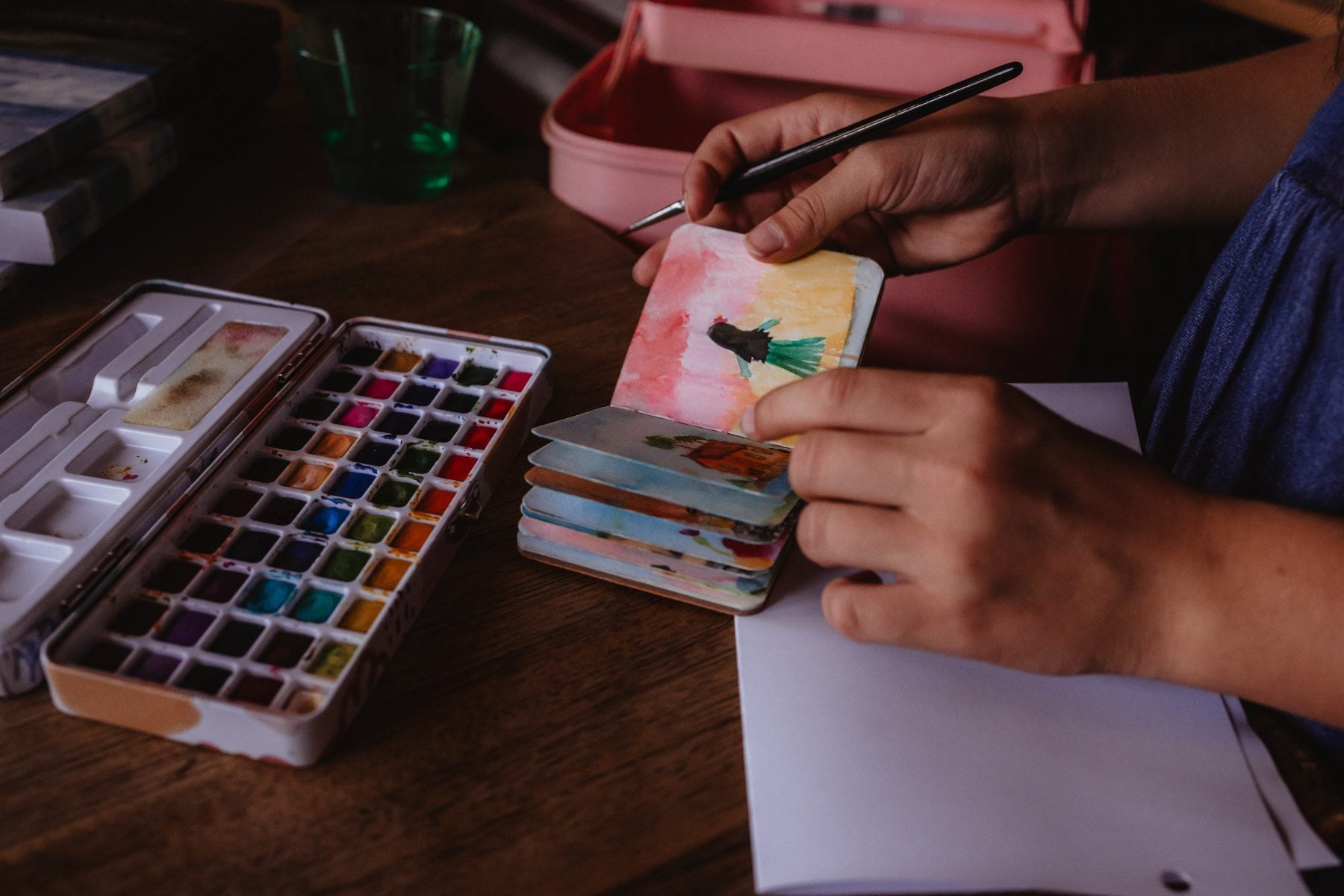 Hands paint a small watercolor artwork at a wooden table