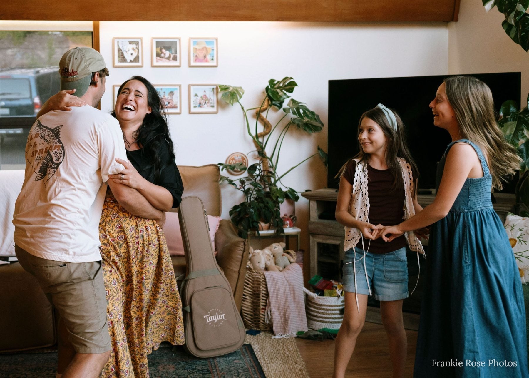Family laughs together in living room beside a ukulele case, two daughters watching