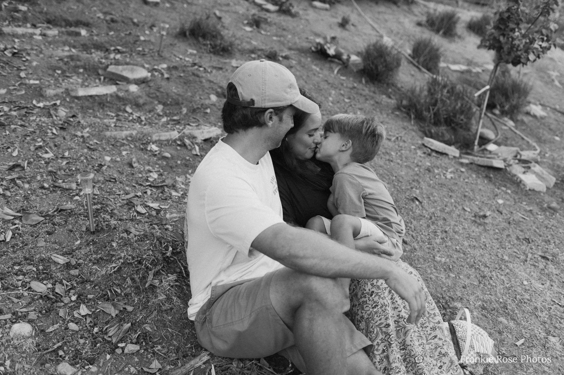 Dad and mom kiss softly with toddler on grassy Lakeside hillside, black and white