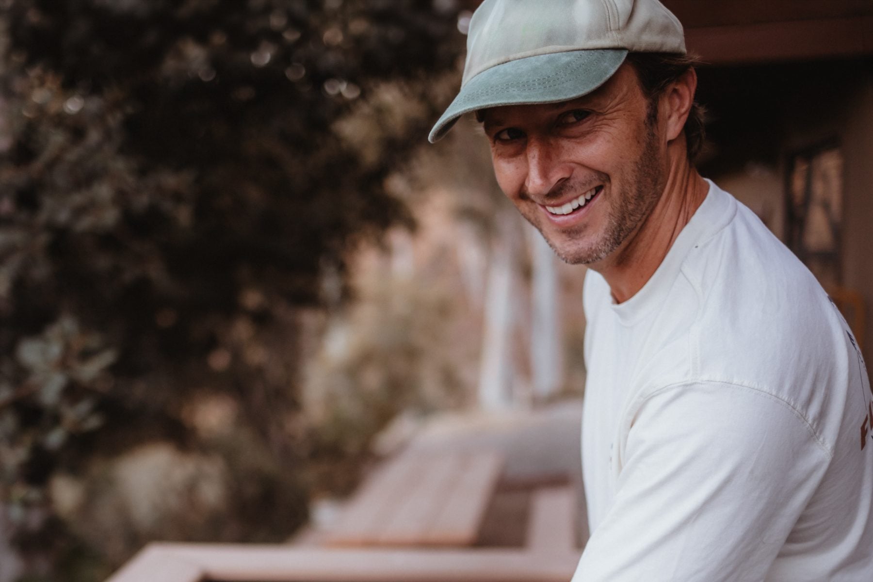 Smiling man in ballcap sits on a wooden deck railing outdoors