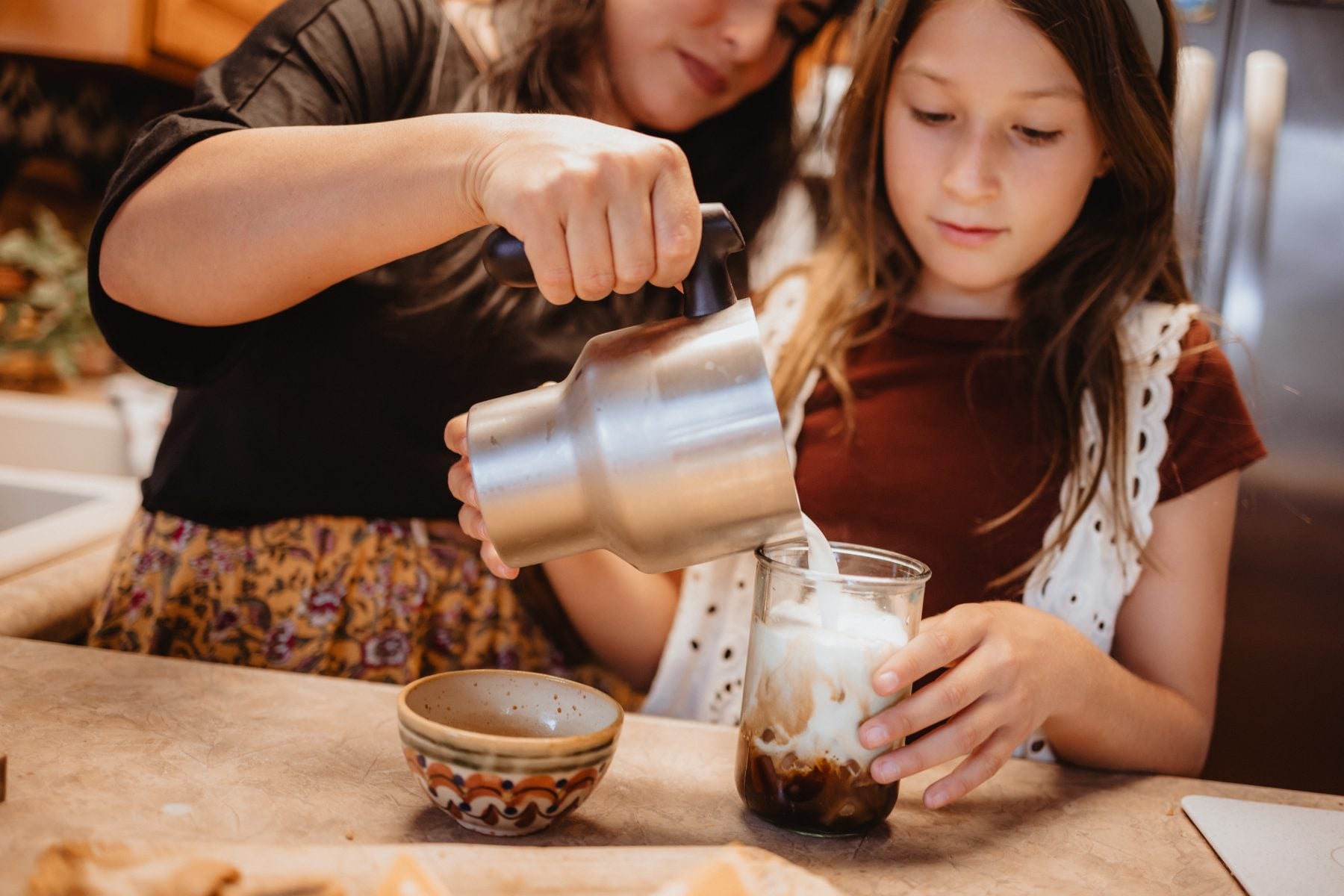 Mom pours steamed milk into iced coffee for daughter in Lakeside kitchen